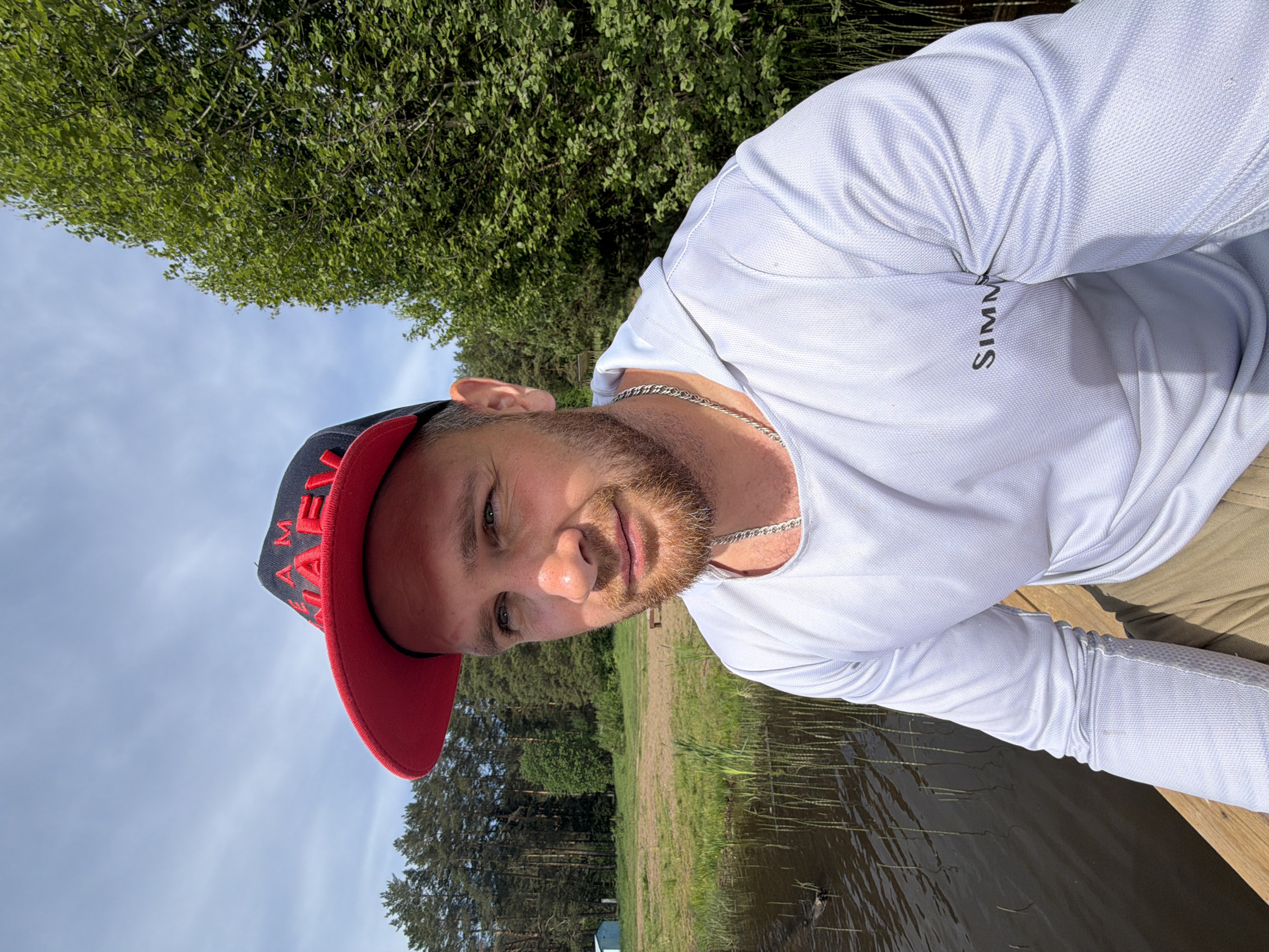 The image is a selfie of a man with a beard and a cap with a red brim. The cap has some lettering on it, partially visible. He is wearing a white long-sleeved shirt with the word "SIMMS" printed on the upper right. A silver chain is visible around his neck.
He appears to be near a lake or body of water with trees in the background. The vegetation suggests it's likely a natural setting. Based on the prompt, the location is Prikamskii, Russia.
