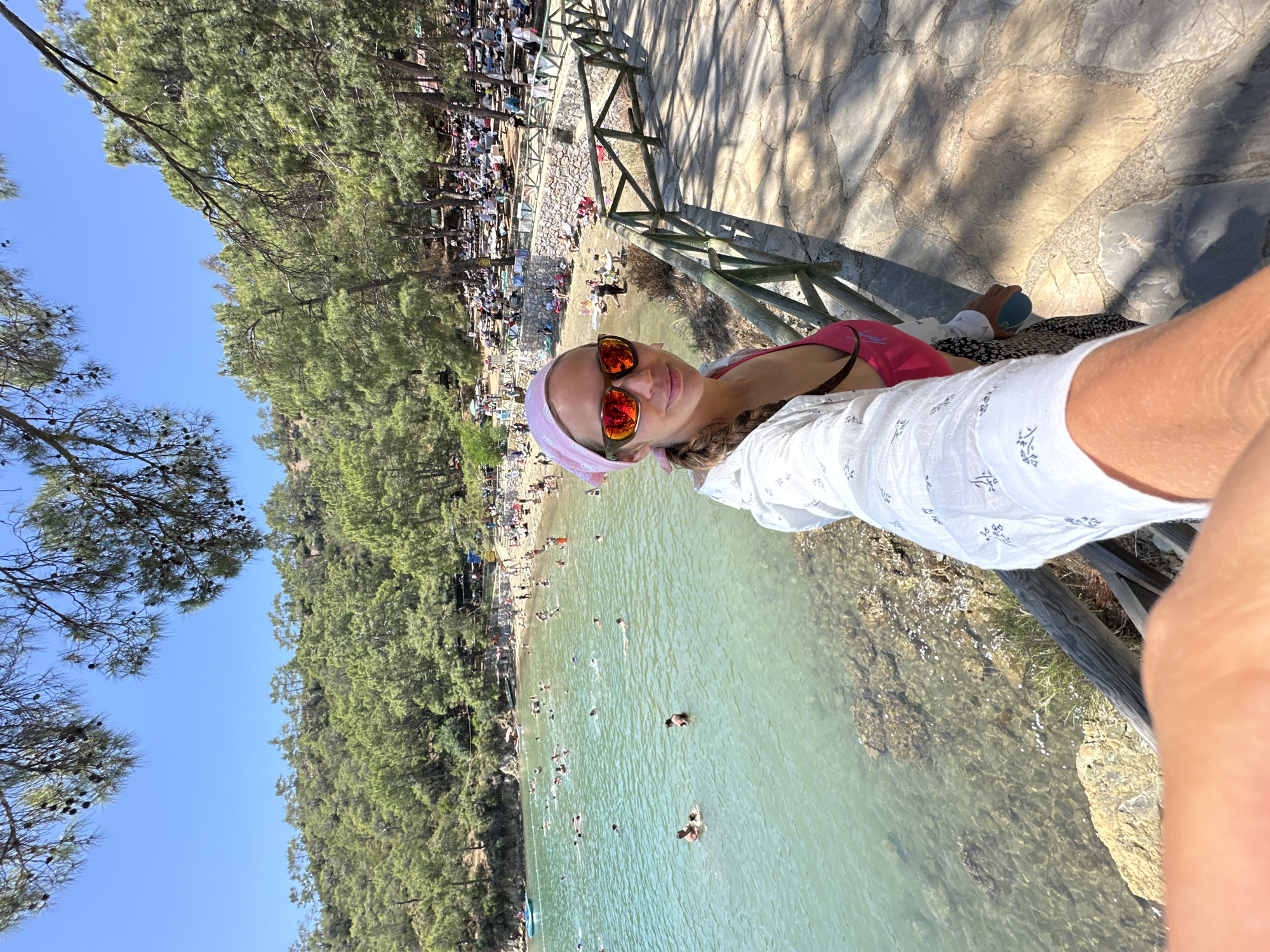 A sideways, eye-level shot shows a woman taking a selfie overlooking a beach area. The woman wears a pink headscarf and sunglasses. In the background, the beach is crowded with people swimming and sunbathing along the shore. A dense forest lines the beach. A wooden fence stands between the photographer and the beach. A tiled walkway is visible on the right. Based on visual clues, this location is likely Ölüdeniz, Fethiye, Turkey.