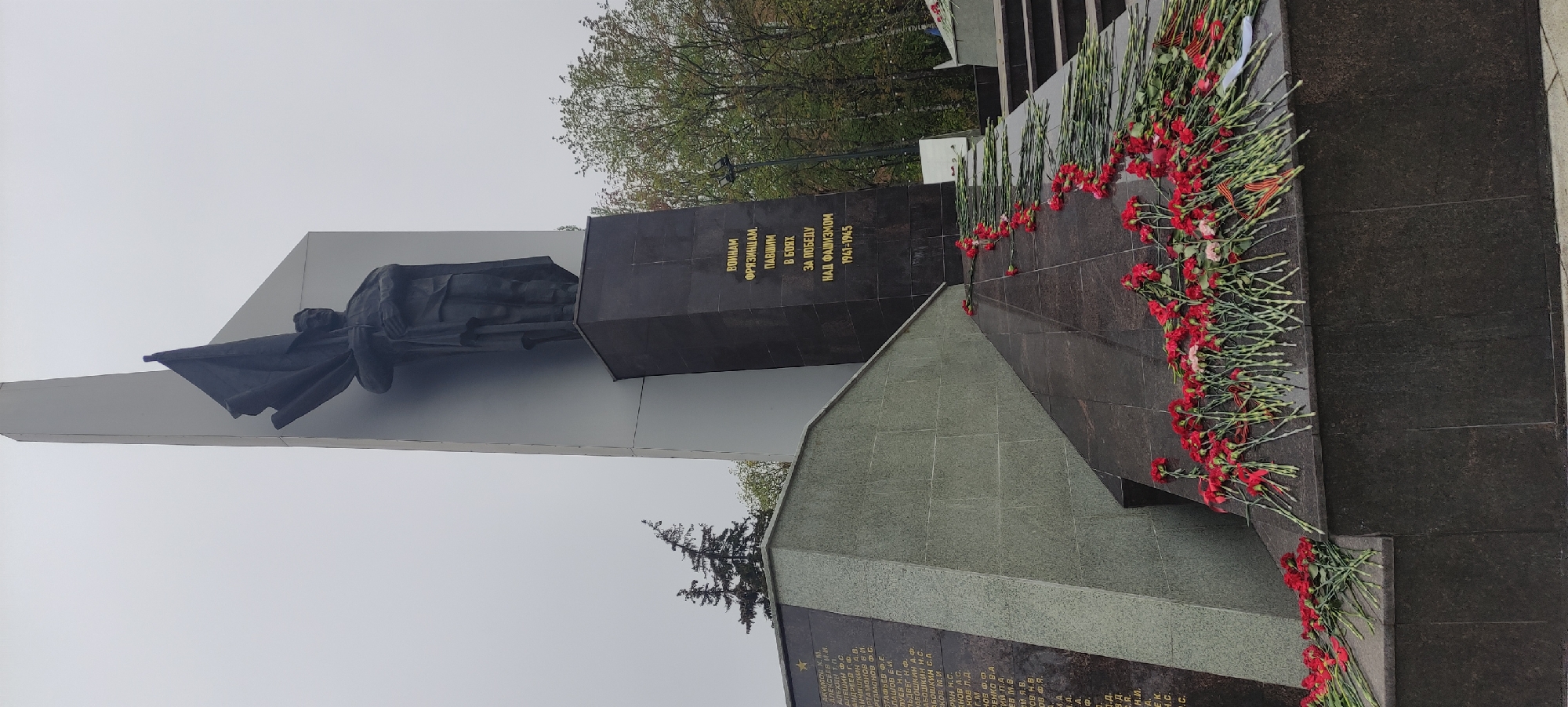 This is a photograph of a war memorial located in the city of Fryazino, Russia. The monument includes a statue of a figure draped in what appears to be a flag, positioned atop a gray, angled structure. Adjoining this is a darker, vertically oriented block inscribed with Cyrillic text in gold lettering. The inscription "ВОИНАМ ФРЯЗИНЦАМ, ПАВШИМ В БОЯХ ЗА ПОБЕДУ НАД ФАШИЗМОМ 1941-1945" translates to "To the soldiers of Fryazino who died in battles for victory over fascism 1941-1945". The base of the memorial has an angled section covered in dark stone, bearing an engraved list of names. Red carnations are laid along the base of the memorial, indicating remembrance. The sky is overcast and gray. Trees are visible in the background.
