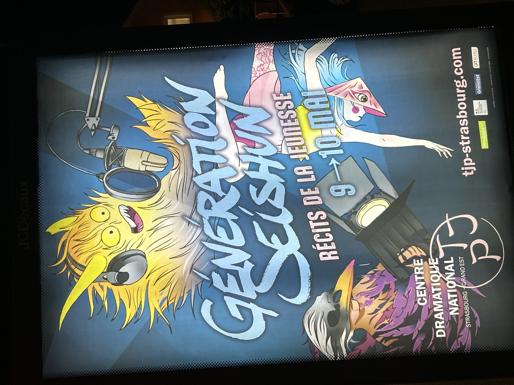 The image shows a poster advertising a theatrical production called "Generation Seishun" (Youth Stories). The poster features cartoon-like figures against a blue background. A yellow, fuzzy creature wearing headphones and a microphone stands to the left. At the top center, a pale female figure with blue hair and a pink triangular headdress is depicted mid-leap. In the bottom right, another figure with purple and gold plumage and a skull-like face is illuminated by a spotlight.
The poster advertises "Récits de la Jeunesse" (Tales of Youth) scheduled for May 9-10. The production is at the Centre Dramatique National (National Drama Center) in Strasbourg, Grand Est, France. The website, tjp-strasbourg.com, is provided, along with logos for the city of Strasbourg, the Grand Est region, and Alsace. The poster also features the JCDecaux logo, indicating that it is likely displayed in a public advertising space.