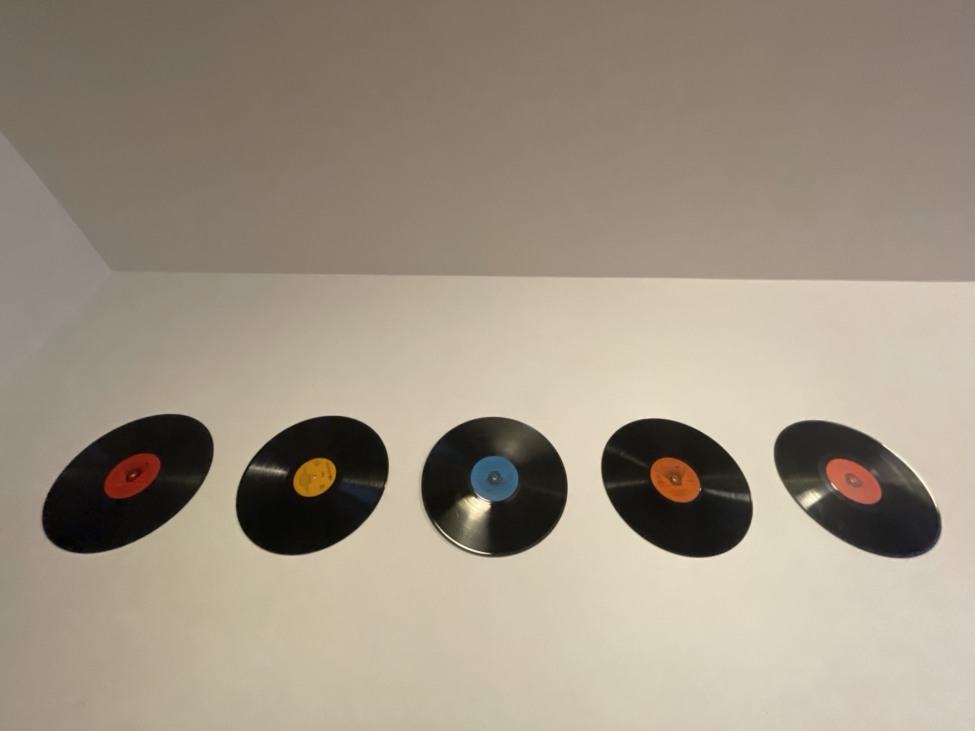 The image shows five black vinyl records mounted on a white wall, with a white ceiling visible above. The records are arranged horizontally, slightly off-center, with varying colored labels: red, yellow, blue, and orange. The environment suggests an interior space. There is no indication of the location being Sofia, Bulgaria, or any other specific city. No people are visible in the image, and there are no discernable actions or interactions taking place.