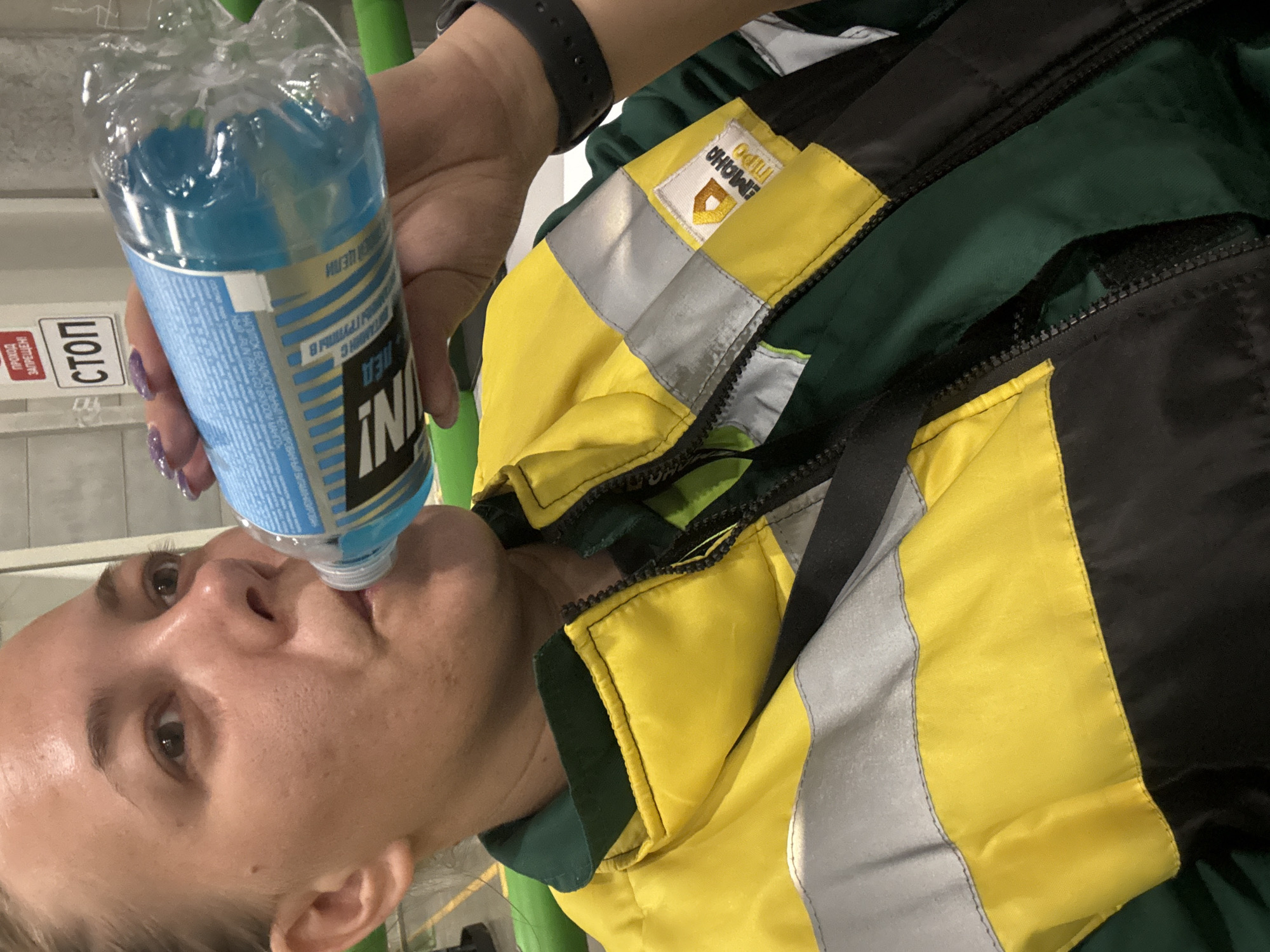 A woman is drinking from a plastic bottle of blue liquid labeled "IN! ЛЕД ВИТАМИН С" and other Russian text. She is wearing a jacket with yellow, black, green, and reflective silver panels, and a black smartwatch on her left wrist. The image seems to be taken indoors, with a sign reading "СТОП" (STOP) visible in the background. The location is not specified as Lupanovo, Russia in the image.