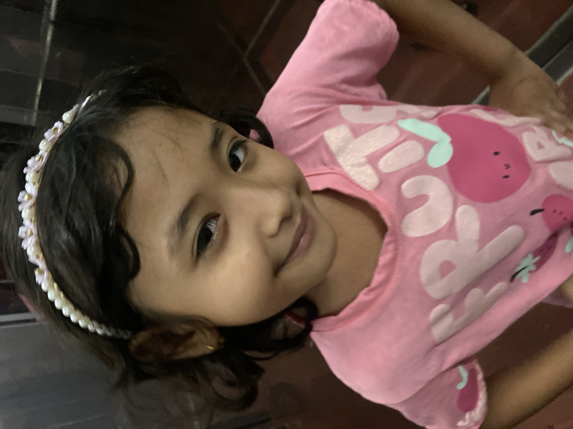 The image captures a young girl, likely of South Asian descent, in an indoor setting in Dhaka, Bangladesh. She is smiling gently and looking directly at the camera with her head tilted to the side. Her dark, wavy hair is adorned with a decorative white headband featuring pearl-like beads and small floral elements, some with gold accents.
She is wearing a bright pink t-shirt with a graphic design on the front. The design includes white letters and a distinct pink fruit, possibly an apple or pear, with a light green leaf. While not fully visible, the text appears to be part of the word "FRUIT" or a similar fruit-related phrase. The background is muted and indistinct, suggesting a casual indoor environment, possibly a home. The lighting is soft, not indicating a specific time of day. The girl's expression conveys happiness and contentment.