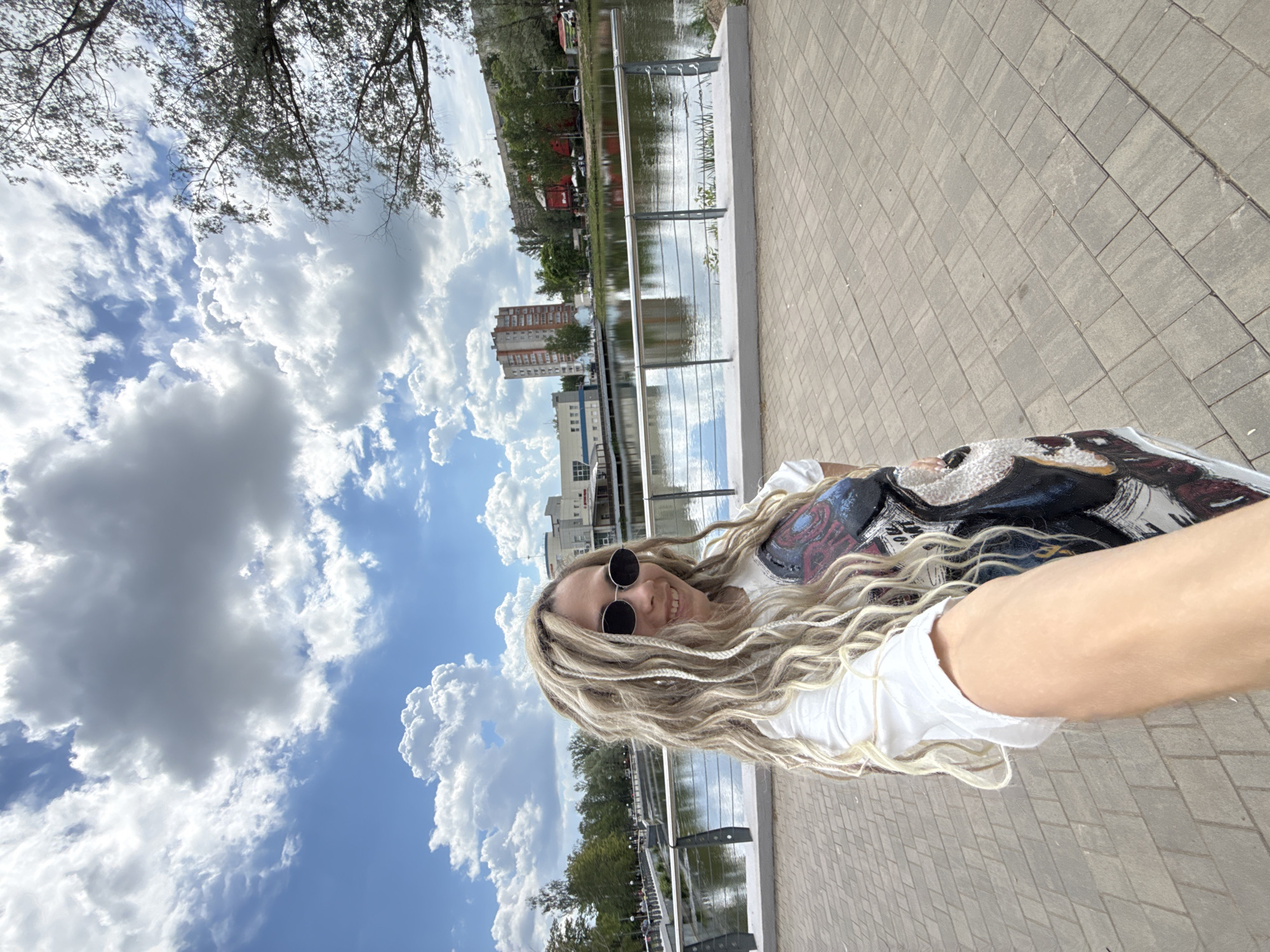 The image is a bright, outdoor selfie, rotated 90 degrees clockwise, featuring a young woman smiling at the camera. She has long, light blonde hair, possibly styled as dreadlocks or braids, flowing over her shoulder. She wears round, dark sunglasses and a white t-shirt, with a colorful, patterned bag or backpack visible on her left side. Her left arm, extended to take the picture, is prominent in the foreground.
The setting appears to be an urban embankment or boardwalk, paved with grey rectangular tiles. To her left, there is a body of water, likely a river or canal, separated by a modern metal railing. The water reflects the bright sky and distant structures. Across the water, an urban landscape unfolds with green trees and multi-story buildings, some appearing residential. The sky is a clear blue with large, scattered white cumulus clouds, indicating a sunny daytime. The overall mood is cheerful and relaxed.
Given the context, the location is Rusanovo, Russia. There is no discernible text visible on clothing or in the background.