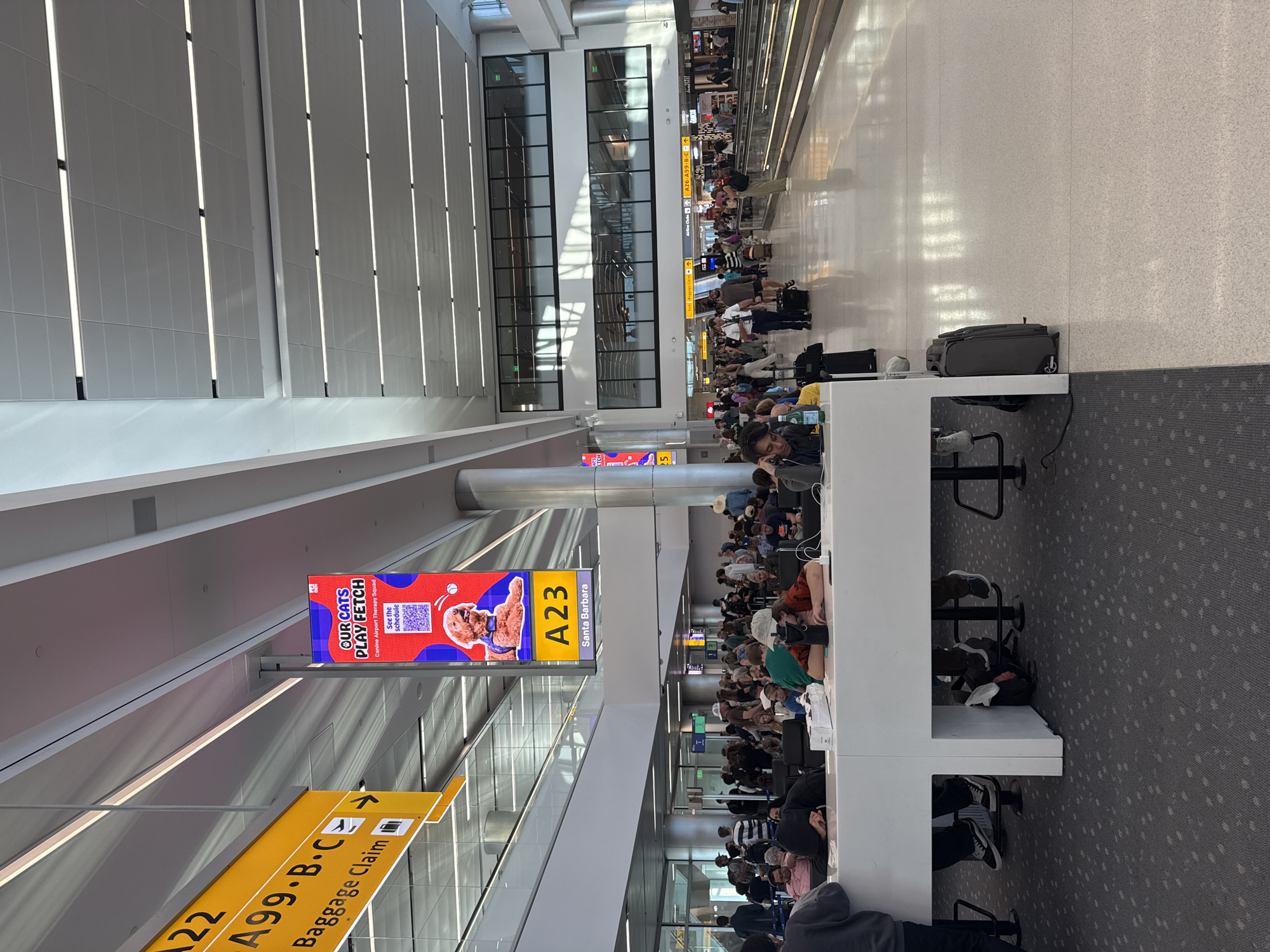 The image shows a busy airport terminal, likely DEN Airport in Denver, United States of America. People are seated at a long white counter, some with their heads down. Luggage is visible on the floor, and signage for "Baggage Claim" and "A23 Santa Barbara" can be seen. A large digital advertisement for "Our Cats Play Fetch" is prominently displayed. The terminal features modern architecture with a high ceiling and ample lighting. A moving walkway is visible in the background, with more people walking along it. The scene depicts a typical airport environment with travelers waiting for their flights or after arriving.