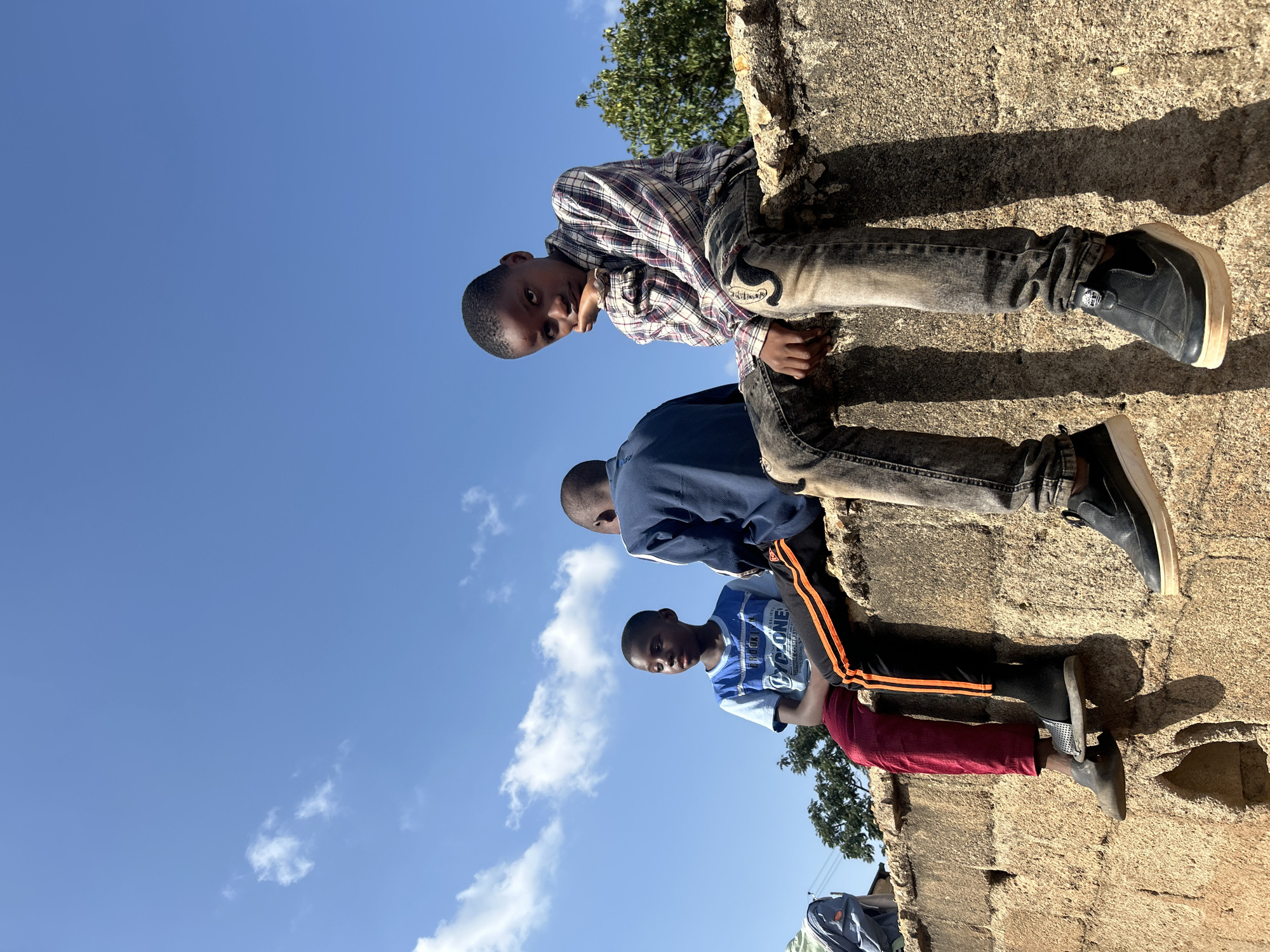 Three young males are seated on a rough, textured concrete or stone wall under a clear, bright blue sky with scattered white clouds. The sun, positioned high to the right, casts long, distinct shadows of the subjects and the wall's irregularities onto its surface. The scene is set outdoors in the city of Jos, Nigeria.
The leftmost male, of apparent adolescent age, is positioned with his legs dangling over the wall's edge, looking towards his right. He wears a light blue and white short-sleeved t-shirt with partially visible text including "BROOKLYN" and "CYCLONE," red trousers over black pants with orange side stripes, black socks, and dark slip-on shoes.
The central male, also an adolescent, is seated with his back to the viewer, facing towards the left. He wears a dark blue long-sleeved top with white stripes on the sleeves, and black pants with orange side stripes are visible on his left leg.
The rightmost male, an adolescent, is facing the viewer, resting his chin on his right hand with his elbow supported by his right knee. He is wearing a plaid long-sleeved shirt with a pattern of red, white, and blue/purple checks, and faded, distressed blue-grey jeans featuring an embroidered patch (possibly an 'S' or stylized logo) on the left knee area. He wears dark slip-on shoes with prominent light-colored soles, his legs also dangling over the wall.
In the background, beyond the wall, green foliage from trees is visible, and faint outlines of distant structures can be perceived under the clear sky. A piece of light-colored fabric is draped over an unseen object on the far left.