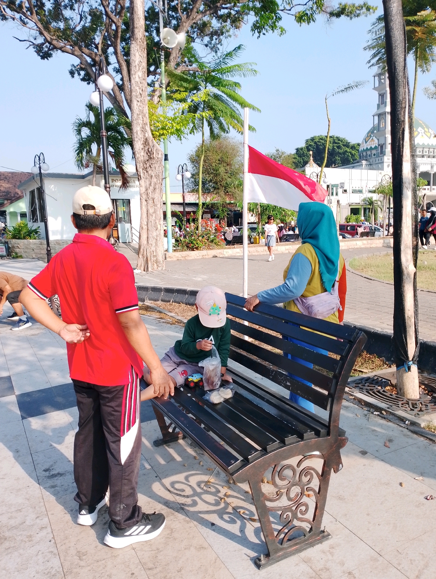 The image depicts a sunny outdoor scene with several people in a park-like area. In the foreground, a person wearing a red shirt and black pants with white and red stripes stands with their back to the camera, facing a black park bench. On the bench, a child wearing a light-colored baseball cap and a green long-sleeved shirt is seated, holding a plastic bag and a toy car. To the right of the child, an adult wearing a teal hijab and a yellow and blue shirt is standing and holding onto the back of the bench.
In the background, a large tree stands prominently, with a flagpole displaying the Indonesian flag. Behind the flag, a white building with a dome and minaret, resembling a mosque, is visible. Palm trees and other foliage are scattered throughout the area. The ground is paved with light-colored tiles and concrete. Several other individuals and parked vehicles can be seen further in the background. The overall impression is a public space in a city, likely a recreational area with cultural elements. Based on the context provided, the location is identified as the city of Probolinggo, Indonesia, specifically in front of the Probolinggo City Hall, which features a mosque with a distinctive dome and minaret.