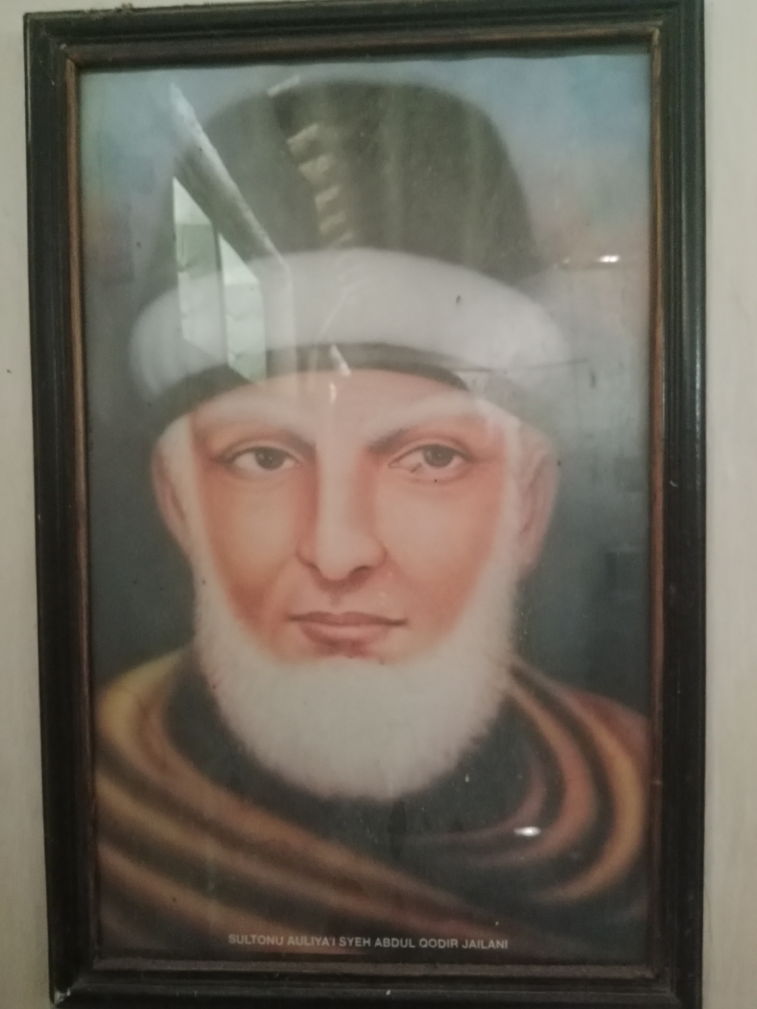A framed portrait of a man with a white beard and a turban is displayed on a wall. The man's face is rendered in warm tones, with his eyes looking directly at the viewer. He is wearing a dark-colored garment that drapes around his shoulders. Below the portrait, a name is inscribed: "SULTONU AULIYA SYEH ABDUL QODIR JAILANI". The portrait appears to be hanging in an indoor space, possibly a room in Ringinrejo, Indonesia, given the context. The image is slightly out of focus and shows some reflections on the glass covering the portrait.