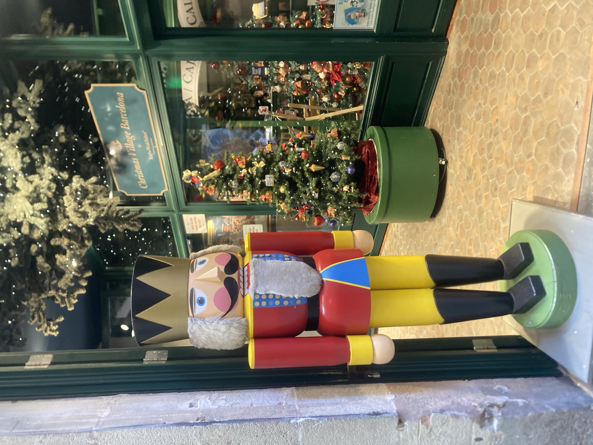 The image depicts the exterior of a Christmas shop in Barcelona, Spain, specifically "Christmas Village Barcelona by Käthe Wohlfahrt". A large, brightly colored nutcracker figure stands prominently on the left side of the frame, facing forward. The nutcracker is dressed in a red coat with yellow accents, yellow pants, and black boots, topped with a golden crown.
Behind the nutcracker, a green doorway leads into the shop, revealing a festive interior. A decorated Christmas tree, adorned with ornaments and lights, is visible through the shop window. The window also displays more Christmas trees and merchandise within the store. A sign above the window reads "Christmas Village Barcelona". The floor in front of the shop is paved with hexagonal tiles. The overall scene suggests a commercial establishment decorated for the Christmas season in a European city.