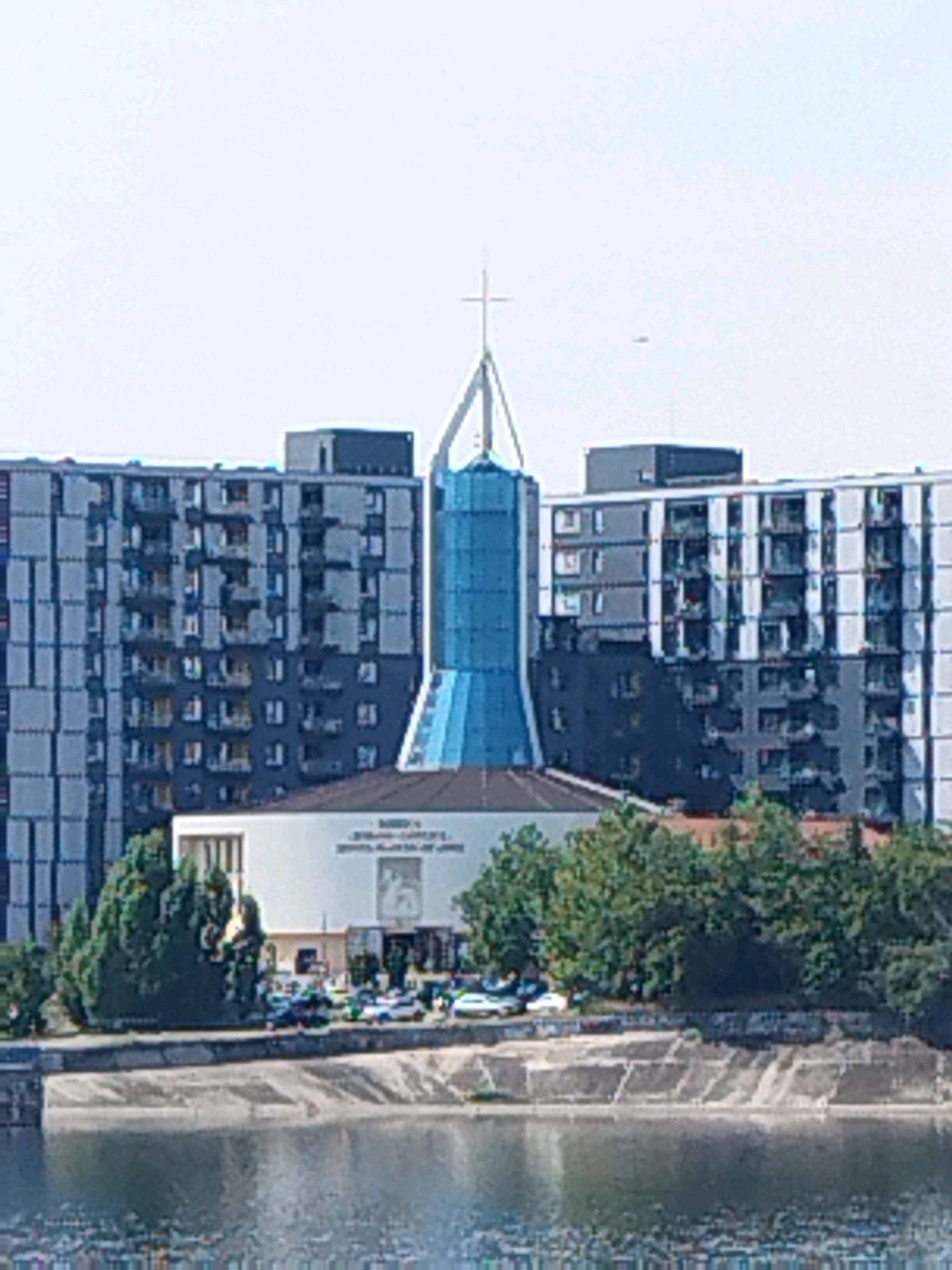 The image shows a contemporary church structure with a distinct blue, glass spire topped with a cross, situated in Bucharest, Romania. The church is flanked by tall, modern apartment buildings with dark facades and numerous windows. In front of the church, a white building with a rectangular entrance and some signage is visible. A row of trees separates the buildings from a concrete embankment, which overlooks a body of water. Several vehicles are parked in a lot in front of the white building. The sky is overcast, and the overall scene depicts an urban environment.