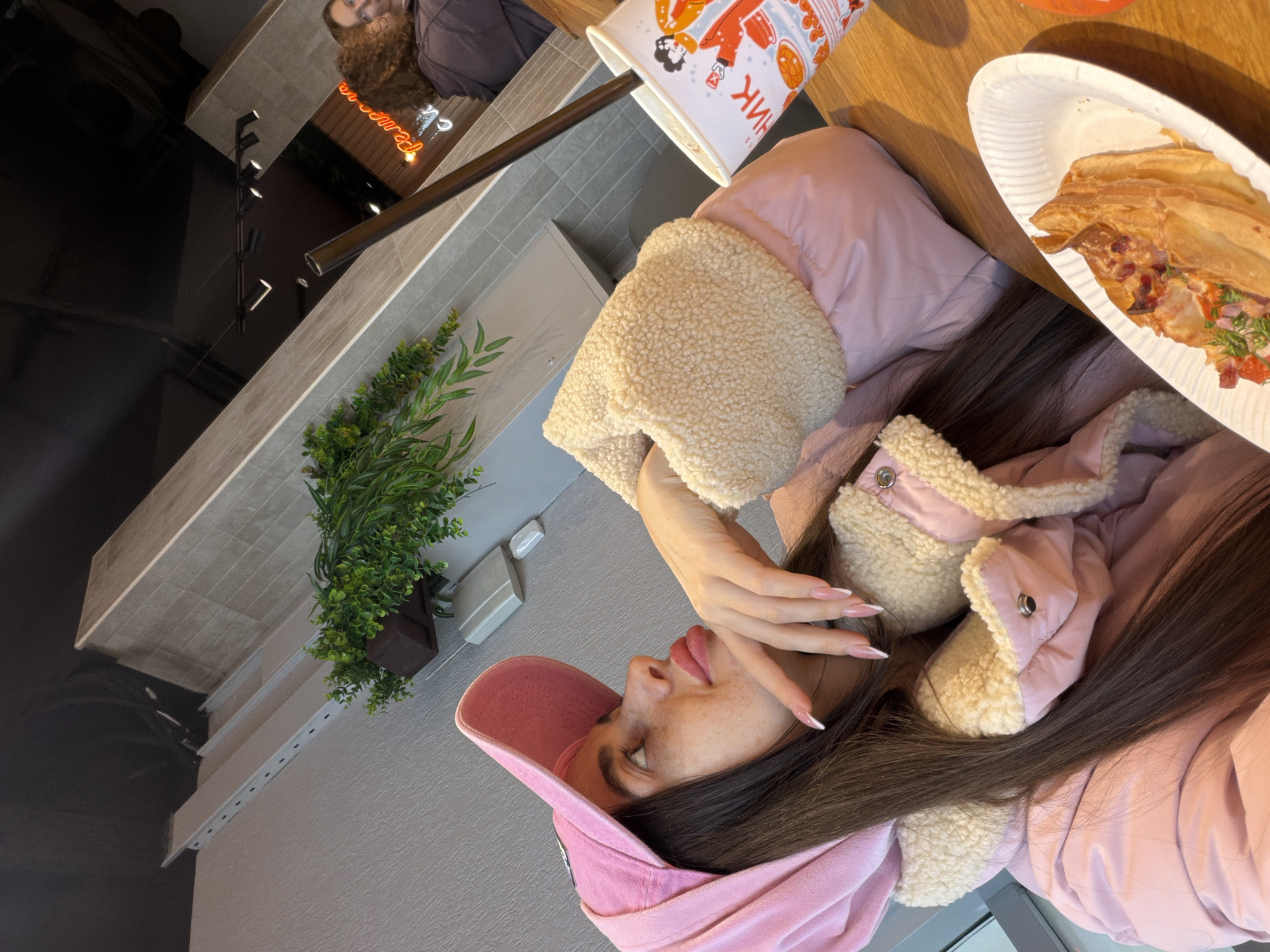 A young woman with long dark hair and visible freckles is seated at a light-colored wooden table inside a cafe, identified as a "Remarka" location in Saint Petersburg, Russia, due to a reflected neon sign displaying the Cyrillic text "Ремарка". She wears a pink puffer jacket with a cream-colored fleece lining and a matching pink baseball cap, with the hood of her jacket partially covering the cap. Her right hand, adorned with French-tipped nails, is positioned near her chin as she looks upwards and to her left with a pensive expression. On the table are a white oval paper plate holding a slice of savory pastry with diced red and green toppings, and a tall white paper cup with colorful stylized graphics and a dark brown straw. The background features light gray textured walls, sections of light-colored rectangular wall tiles, artificial green plants in a brown planter, and a dark ceiling with track lighting fixtures. A partial reflection of another individual with curly brown hair is visible in the upper right.