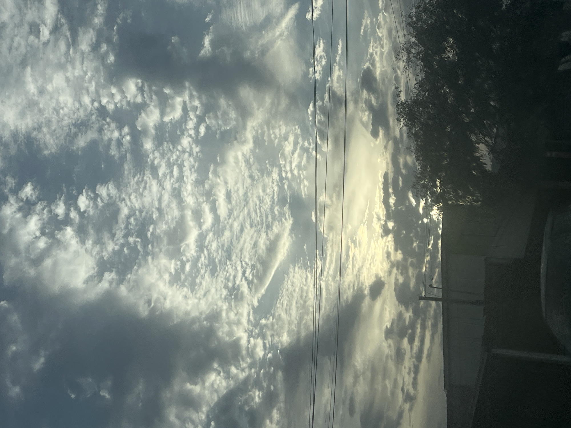 The image depicts a view of the sky with cumulus clouds, possibly taken from a moving vehicle in Arica, Chile. Power lines are visible against the cloudy backdrop. A building and tree are partly visible on the right side of the image. The lighting suggests it is daytime. The exact location in Arica is difficult to determine without more context.