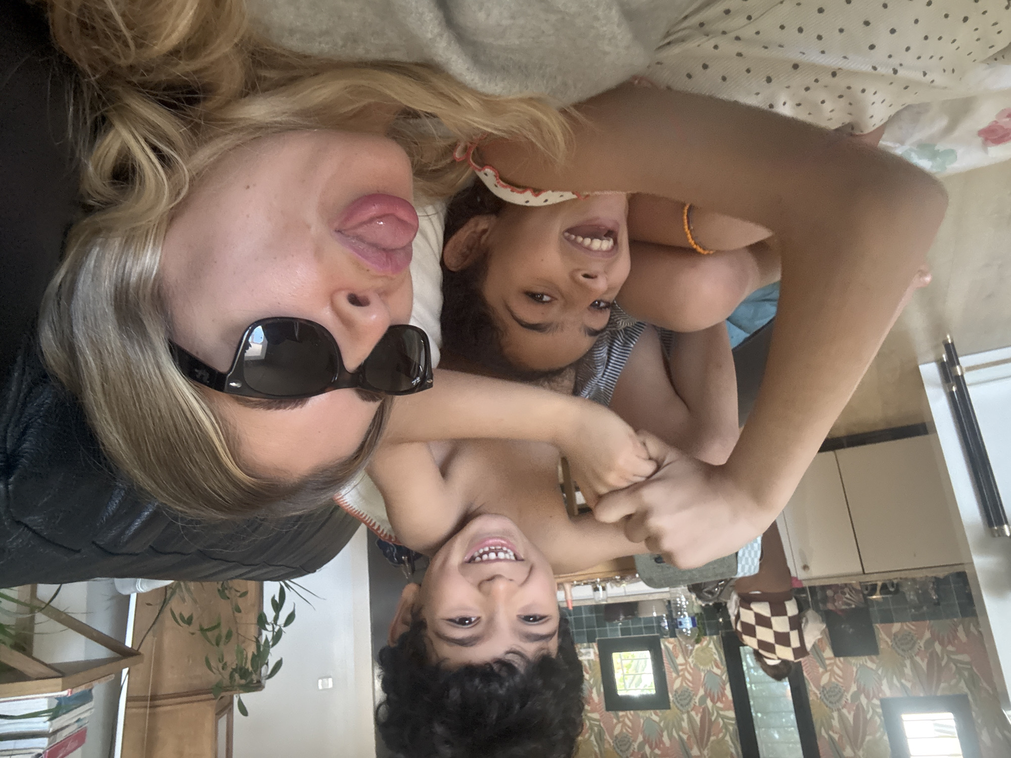 An indoor scene in Rousset, France, captures three individuals in a playful, upside-down perspective. The primary subjects are a blonde woman and two young children, a girl and a boy, all appearing to be lying down or leaning back. The woman, with long blonde hair, is visible on the left, wearing dark sunglasses and a black garment, playfully sticking her tongue out. To her right, a young girl with dark hair smiles broadly, wearing a striped top and an orange or yellow bracelet on her left wrist. Below them, a young boy with dark curly hair is also smiling widely, appearing bare-chested.
The activity taking place is a joyful and casual pose for a photograph, highlighting familial warmth and happiness. Their expressions convey amusement and contentment.
In the background, various domestic elements are visible. Directly behind the subjects, there's a light-colored, possibly polka-dotted, fabric item, alongside a patterned fabric with floral or leafy designs, and a plain light-grey texture, likely indicating bedding or a couch. Further into the background, the setting appears to be a home, possibly blending living and kitchen areas. Visible objects include shelves with books, a plant, a mirror, white kitchen cabinets, and a wall adorned with a patterned wallpaper featuring leaves or flowers. A checkered item and a water bottle are also present. The lighting suggests it is daytime. No discernible text is visible on any object.