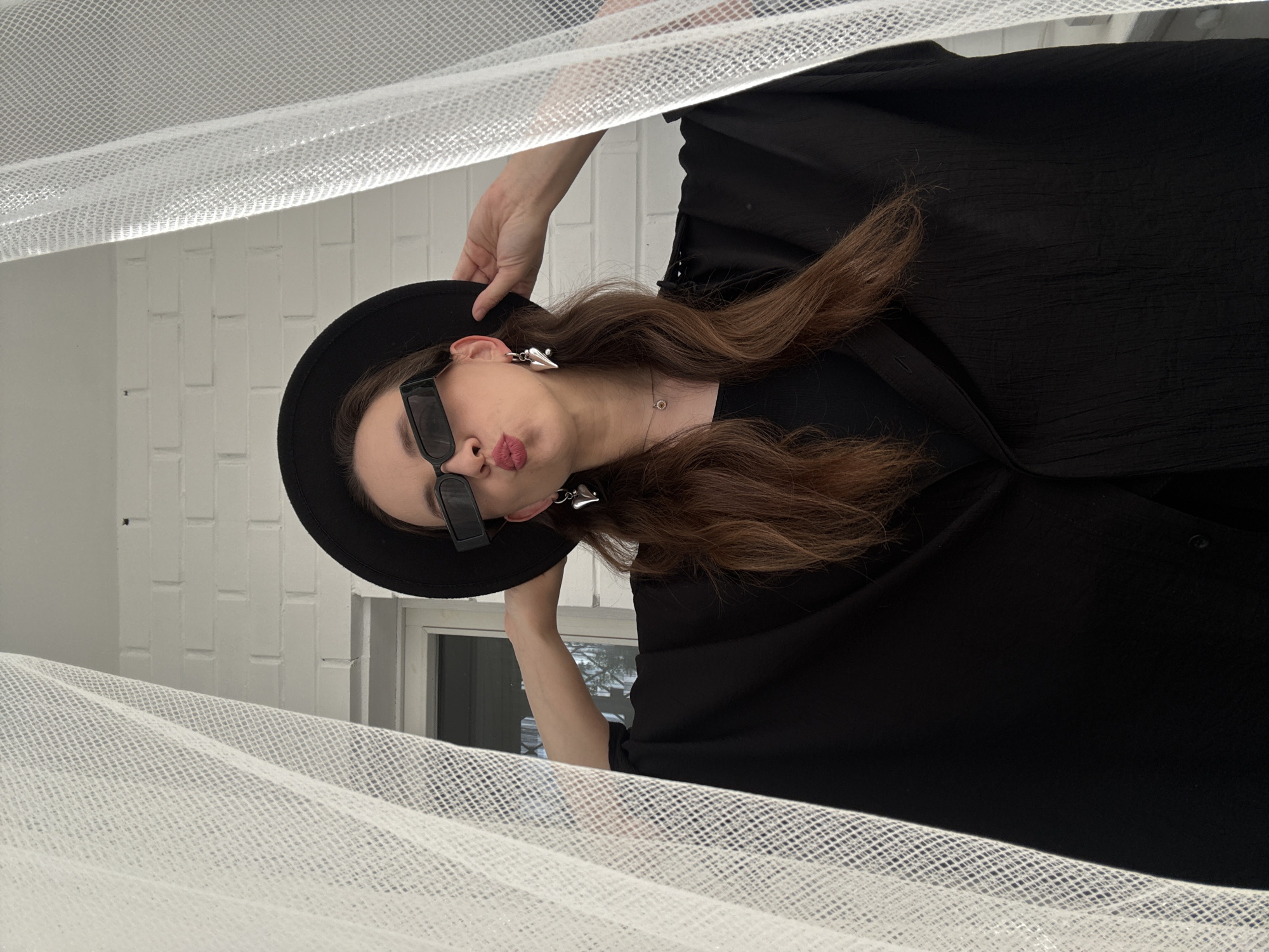 A woman with long, brown hair, wearing a black hat and sunglasses, poses in front of a white brick wall and a window. She is wearing large, silver earrings and a black outfit. A white, sheer fabric is draped in the foreground. The location is Chaikovsky, Russia.