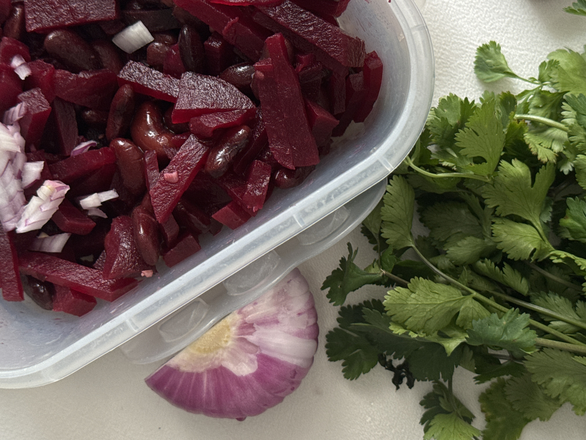 The image shows a plastic container filled with diced beets, kidney beans, and chopped white onion. Next to the container, a half-cut red onion sits alongside a bunch of fresh cilantro. The scene is situated on a plain white surface. There is no evidence to suggest a link to the city of Akulovo or Russia.