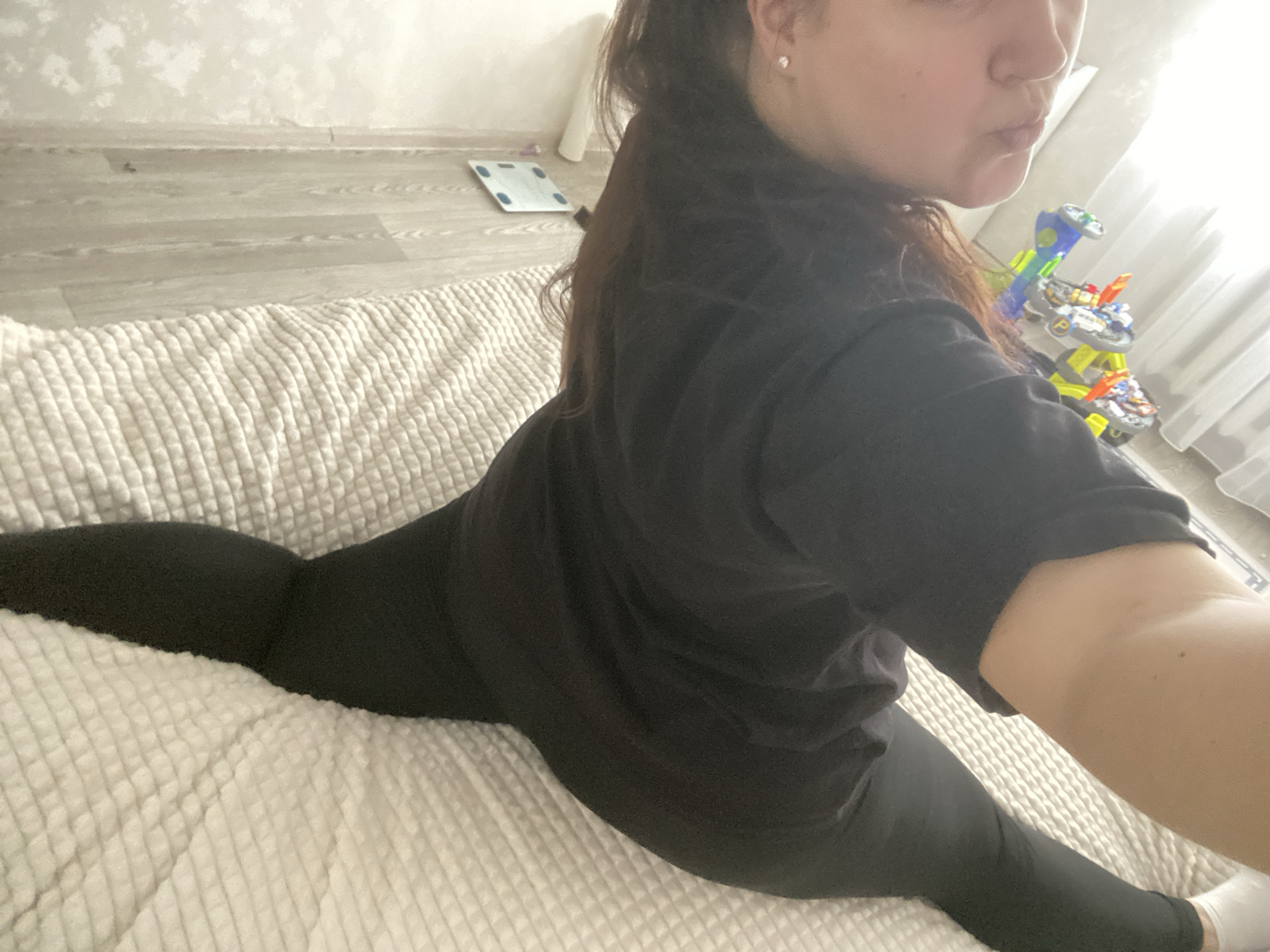 A woman in black leggings and a black t-shirt is performing a split on a white textured surface, possibly a bed or mattress. The background shows a wall with a scale and a toy. The location is unknown and there is no way to determine if the location is Sochi, Russia or any other place.