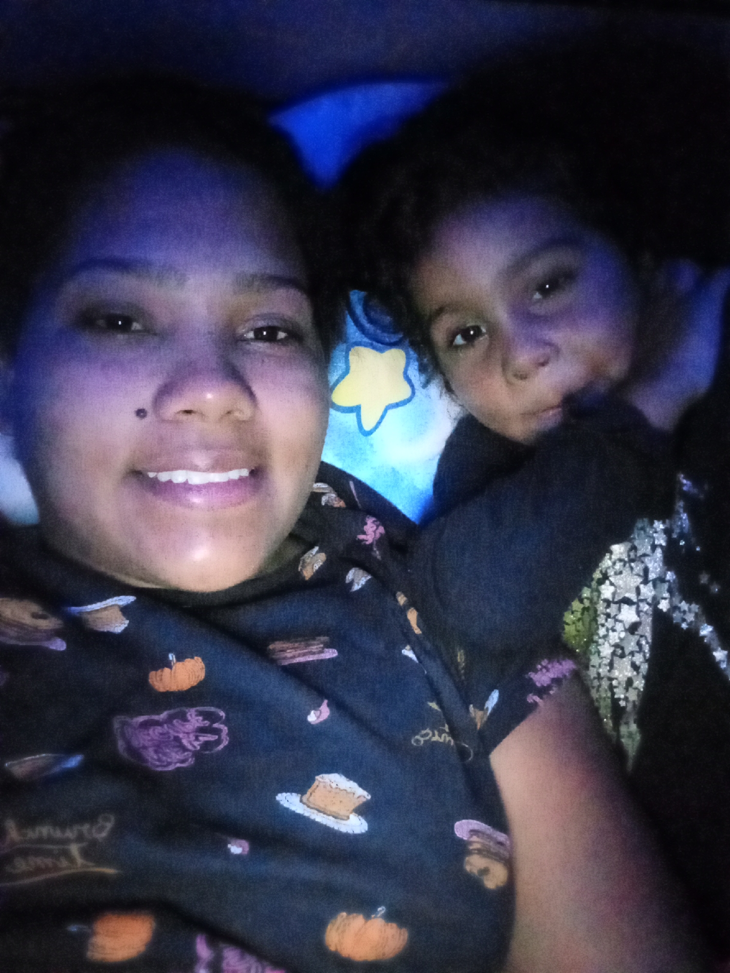 The image shows a selfie taken at night, likely indoors, with a woman and a child lying down together. The woman is smiling, facing the camera directly. She has dark hair and a mole near her nose. Her top is dark with images of pumpkins and other fall-themed items printed on it. The child is lying to her right, also looking at the camera, with their hand near their face. The child's clothing appears to have star-shaped glitter on it. A pillow with a yellow star is visible behind them. The lighting is dim and blue-toned. There is no information in the image to suggest that it was taken in Parroquia Ocumare, Venezuela.
