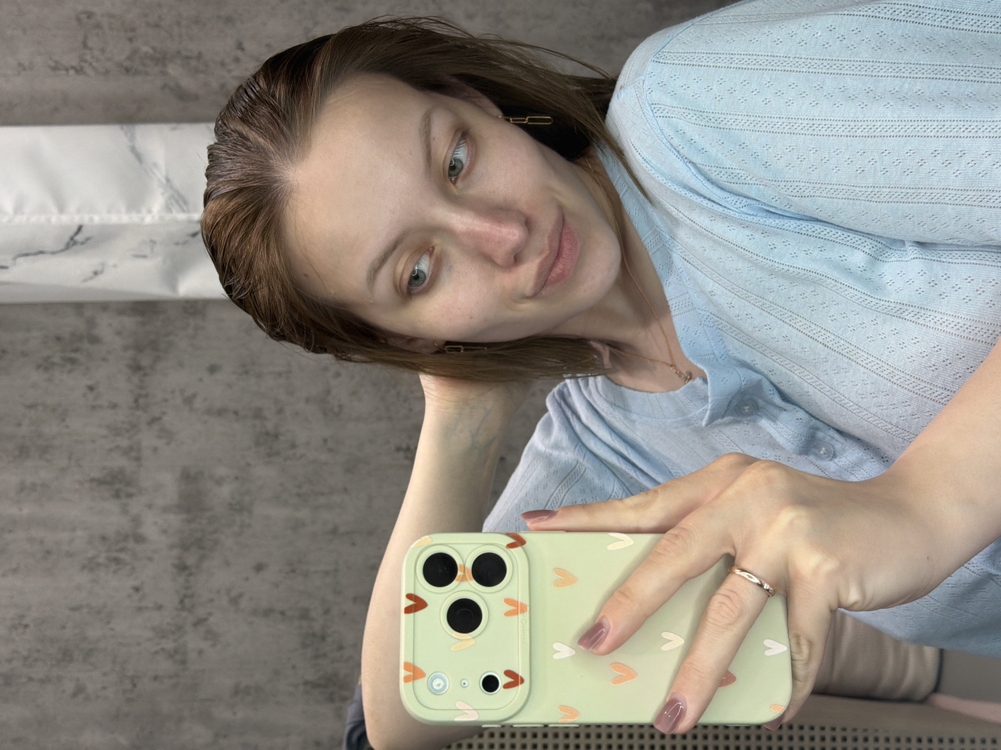 A woman is holding a light green iPhone with a heart-patterned case, taking a selfie. She is wearing a light blue, short-sleeved, textured top and gold chain earrings. Her hair is brown and appears wet. She is wearing a gold ring on her ring finger. The background appears to be a gray wall and a section of white marble-patterned material. There is no indication that the image was taken in Reshetnikovo, Russia.