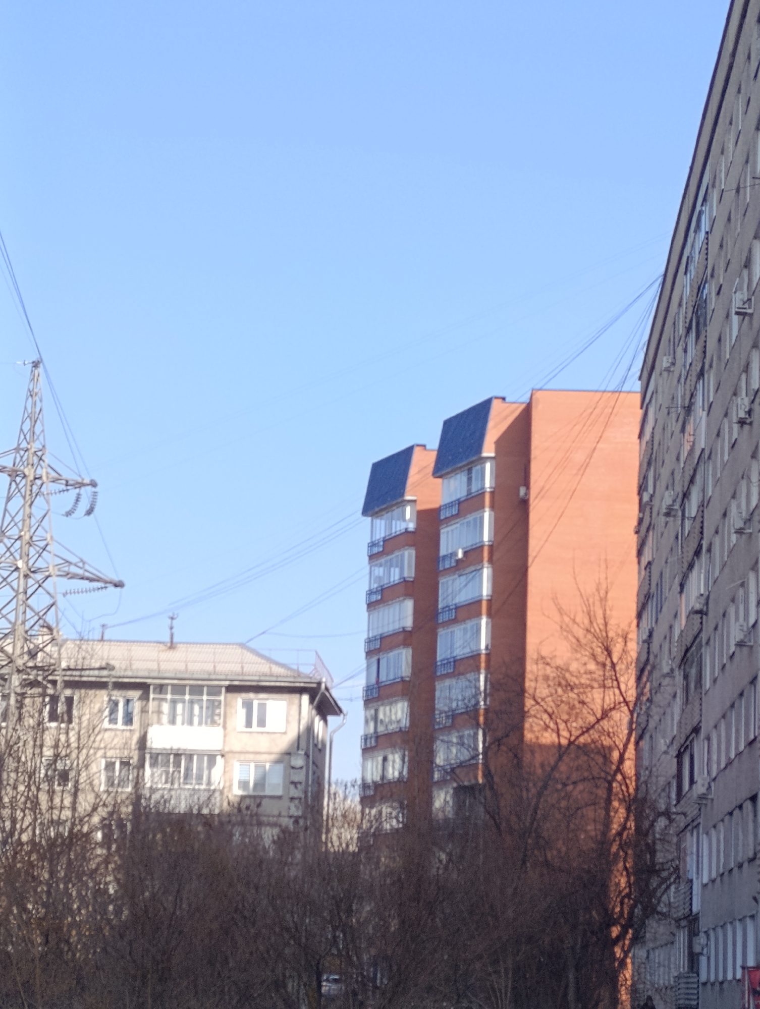 The image shows a street scene in Krasnoyarsk, Russia, with a mix of residential buildings. The buildings vary in age and style. On the left is a power transmission tower. Overhead power lines connect the tower to the buildings, and connect the buildings to each other. The foreground is obscured by trees without foliage. The sky is clear and blue.