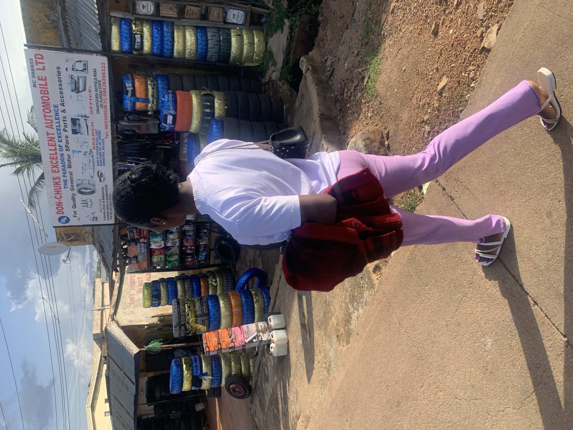 This image shows a young woman from the chest down, wearing a white long-sleeved shirt, light purple pants, and white strappy sandals. She is holding a red sweater or scarf draped over her arm. She is standing on a paved surface next to a shop that sells automotive spare parts, specifically tires.
The shop displays many tires stacked on shelves and on the ground. A prominent sign for "DON-CHUKS EXCELLENT AUTOMOBILE LTD" is visible, listing the company as "THE PARAGON OF EXCELLENCE" and specializing in "Quality General Motor Spare Parts & Accessories." The sign also provides contact information and addresses for their Head Office and Branch, indicating locations in Jos, Nigeria. Other items visible include what appear to be oil or chemical containers and other assorted automotive parts.
The setting is outdoors, under a partly cloudy sky, suggesting daytime. The ground is a mix of paved concrete and dirt with some vegetation. The overall impression is a bustling street-side vendor in an urban area, likely a market or commercial district in Jos, Nigeria, as indicated by the text on the sign. There is no discernible emotion from the visible parts of the subject, but the scene depicts a commercial transaction or a stop at a shop.