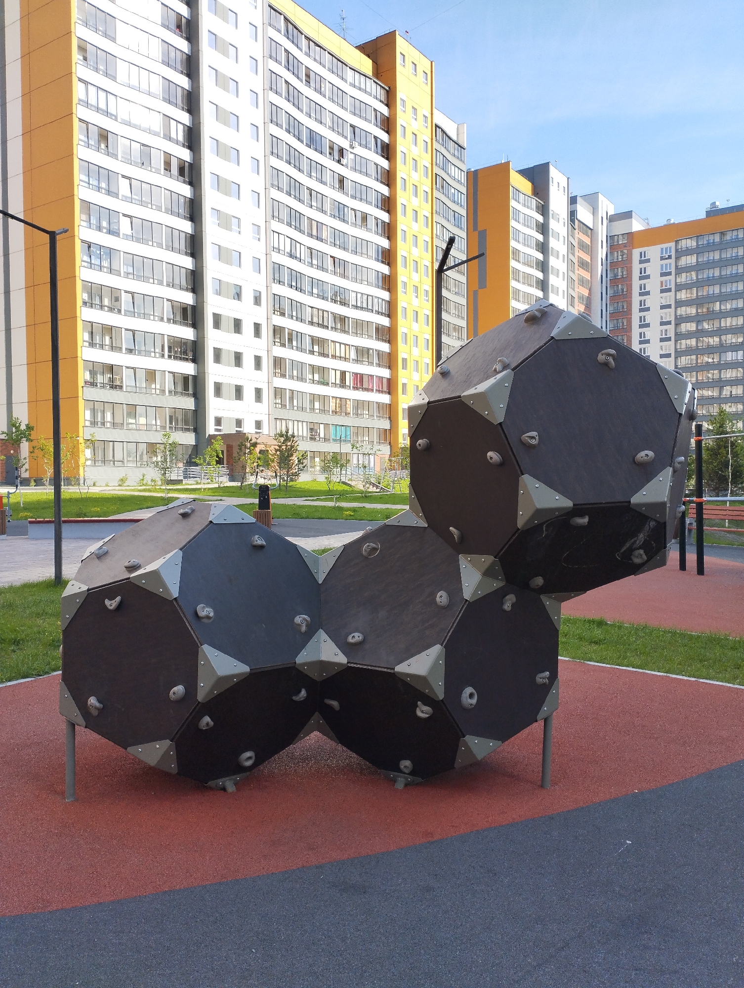 The image shows a playground in Sadovyy, Russia, featuring a unique climbing structure made of interconnected, dark-colored geometric shapes with climbing holds attached. The structure sits on a cushioned, red and dark gray surface surrounded by green grass. In the background, tall residential buildings with a mix of white, gray, and orange facades rise against a clear blue sky. A light pole stands to the left of the climbing structure. A bench and exercise bars are visible further back in the playground area.