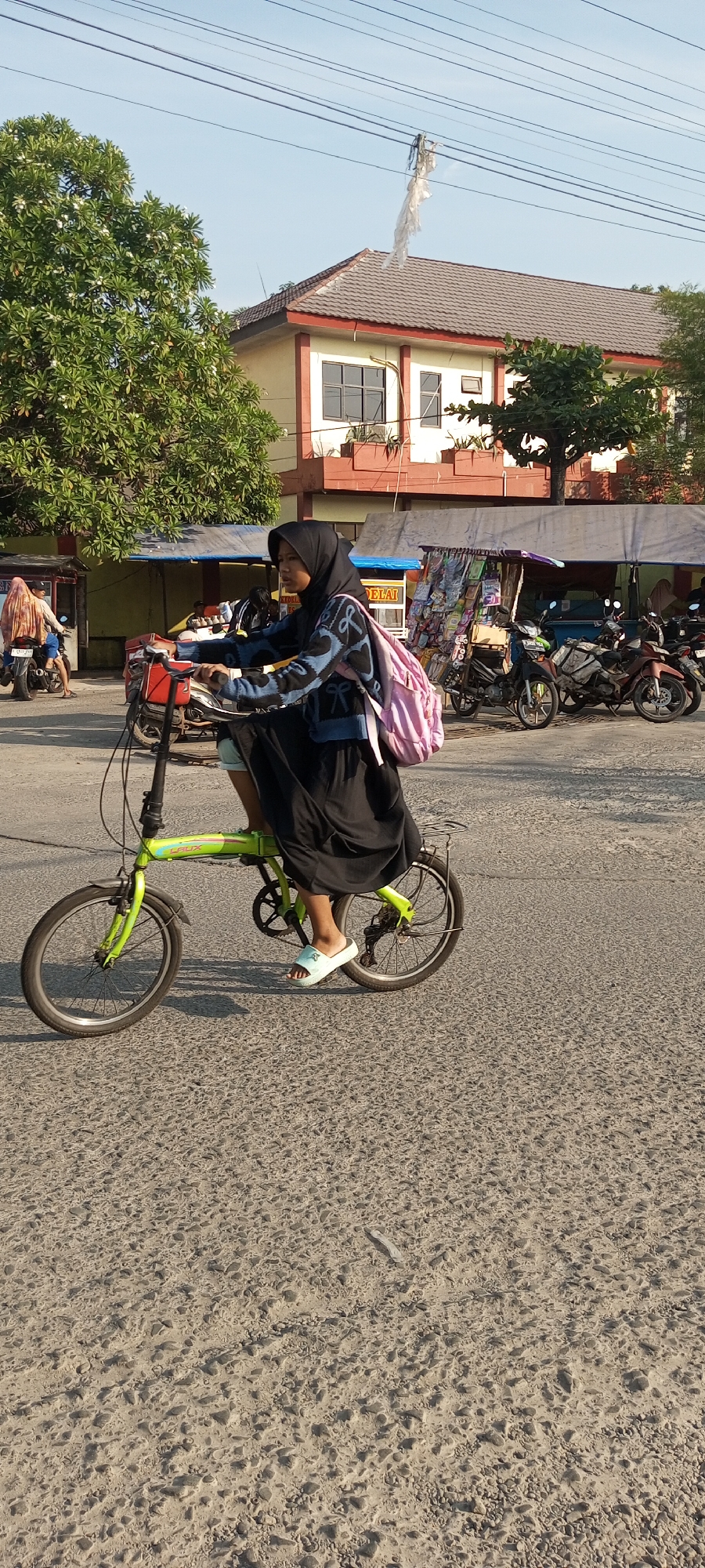 A young woman wearing a black hijab and a blue and black patterned sweater rides a lime green folding bicycle down a paved street in Setu, Indonesia. She has a light pink backpack on her back. She is wearing light blue sandals. The street appears to be made of asphalt with small stones embedded in it. In the background, a two-story building with a brown roof is visible. There are also a few trees with green leaves and some parked motorcycles. In the foreground, there are electrical wires with a piece of white plastic caught on them.