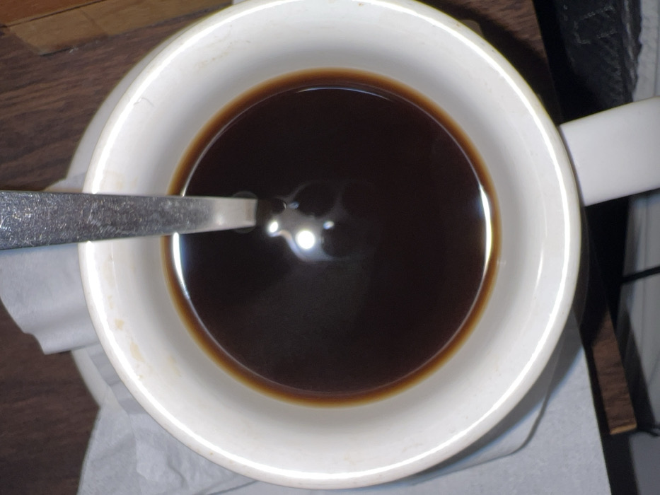 The image displays a white ceramic mug, viewed from an overhead perspective, filled with a dark brown liquid, consistent with coffee. A silver-toned metal spoon is positioned vertically within the liquid, with a distinct circular light reflection visible on the liquid's surface. The inner rim of the mug shows minor brown discoloration. The mug is resting on a surface that includes a crumpled white paper napkin to the left and a dark brown wooden surface, likely a table or countertop, to the right. No individuals are present. The environment is an indoor setting. This scene is situated in Toyapakeh, Indonesia.