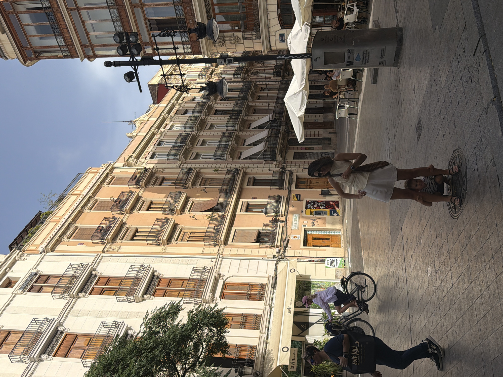 This is a vertical, low-angle shot of a European city street scene. The perspective is looking up at ornate, multi-story buildings with balconies and large windows. The buildings are painted in shades of cream and peach, with dark wrought iron railings. A bright blue sky is visible between the structures.
In the foreground, on the right side of the frame, two women are partially visible. One woman in a white dress appears to be holding a child. The other woman is wearing sunglasses and a dark t-shirt. In the middle ground, a person is bent over a bicycle, possibly adjusting it or preparing to ride. Further down the street, another person is seen cycling.
The street is paved with stone tiles in a diamond pattern. A large metal trash receptacle with some signage is positioned on the sidewalk. Several white chairs are arranged on the patio of an establishment. Faint sunlight suggests it is daytime, likely mid-morning or afternoon.
Visible text includes: "lavin", "lavin snack&bar", "sole piel", "ATM", "RENT A BIKE", "CAU DE SERRANS", and names of local districts or landmarks like "Serrans" and "Furs". The text "valencia" is also present. The overall impression is a lively urban environment.