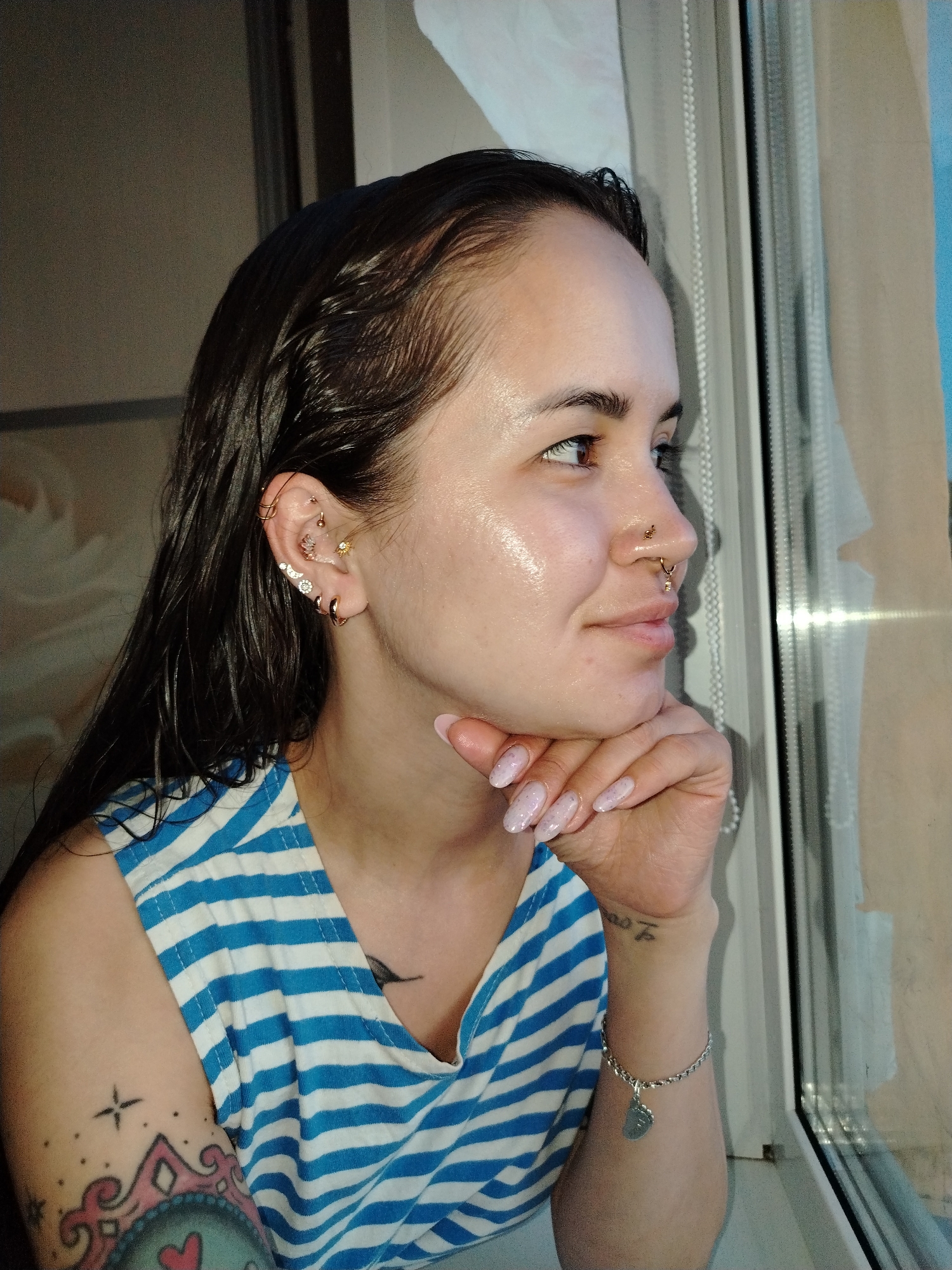 A medium shot captures a woman gazing out of a window. She has dark, slicked-back hair and multiple piercings in her left ear and nose. Her skin is dewy, and she is wearing a blue and white striped tank top. Her left arm, adorned with tattoos, rests against her cheek. A silver bracelet is visible on her left wrist. The background shows a window frame with a translucent curtain. The location is Noviy, Russia.