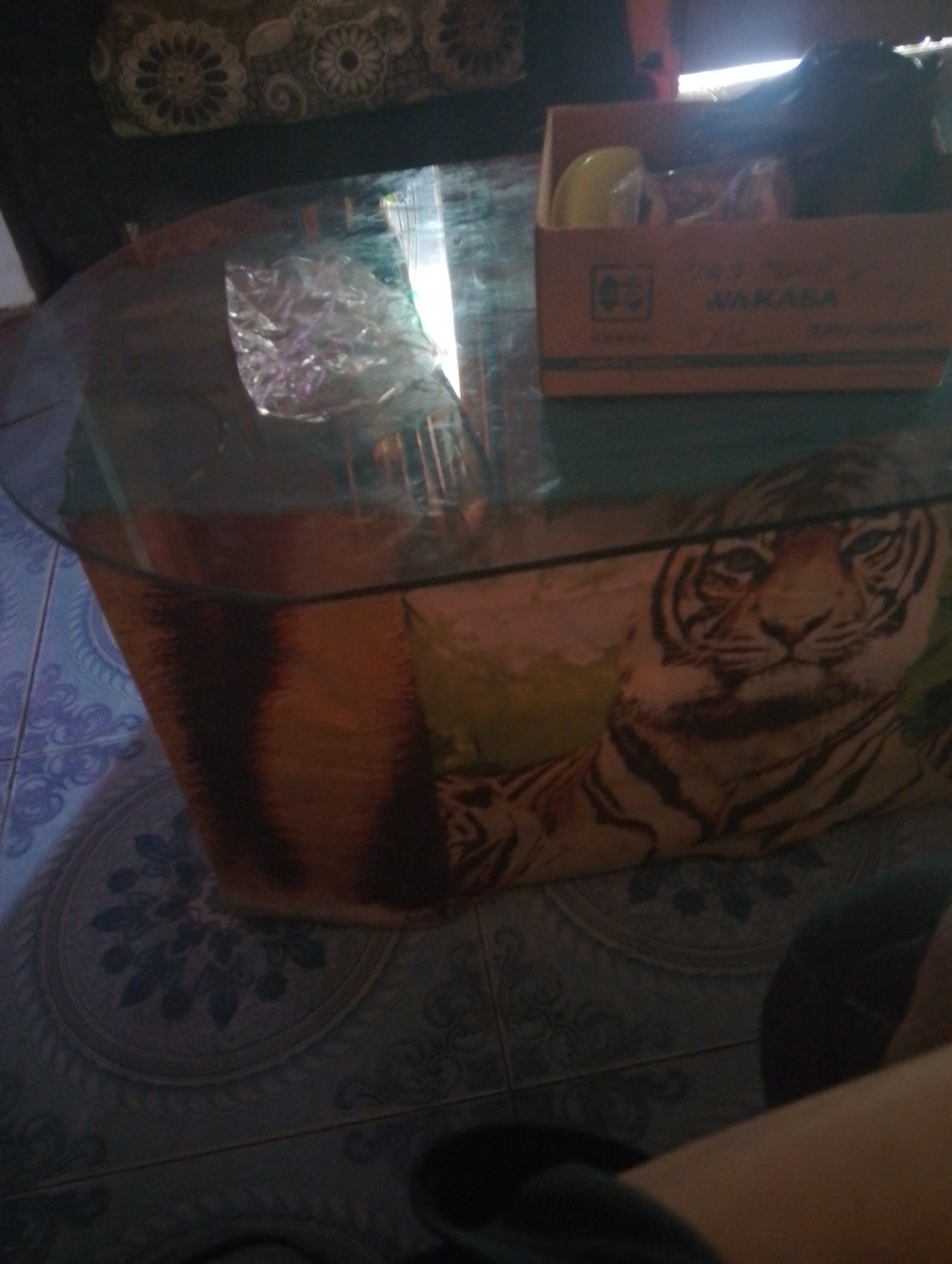 A glass-top coffee table with a tiger-themed base is set on a tiled floor. A cardboard box, labeled "WAKASA," is on the table. The room appears to be dimly lit, and a portion of a patterned couch or chair is visible in the upper left corner. The environment suggests an indoor domestic setting, possibly in Purwakarta, Indonesia, given the context.