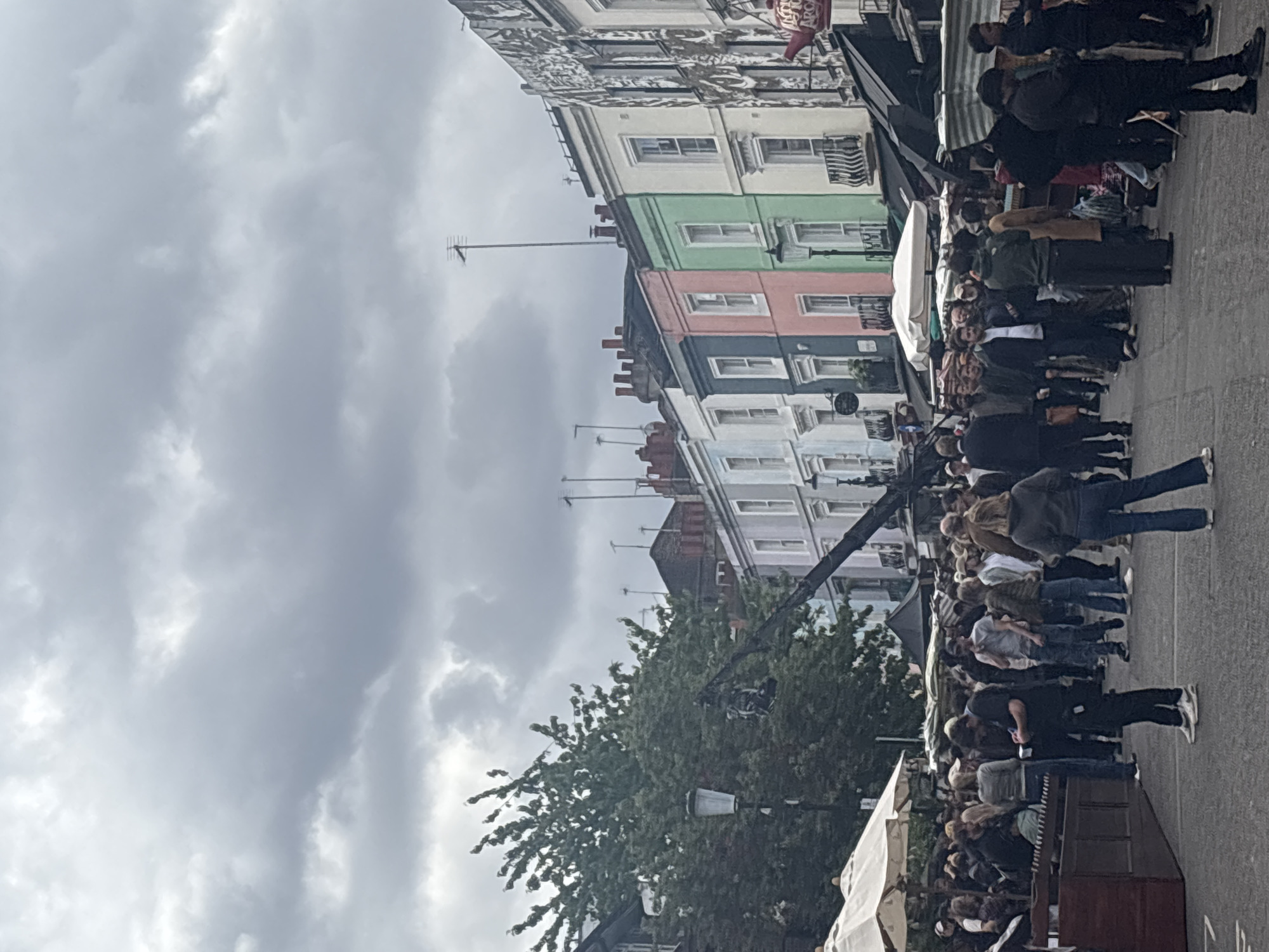 The image depicts a street scene in Notting Hill, London, United Kingdom. A crowd of people is gathered on a paved street alongside colorful terraced houses. The sky is overcast. Several market stalls with white canopies line the street. A black camera boom is visible above the crowd.
