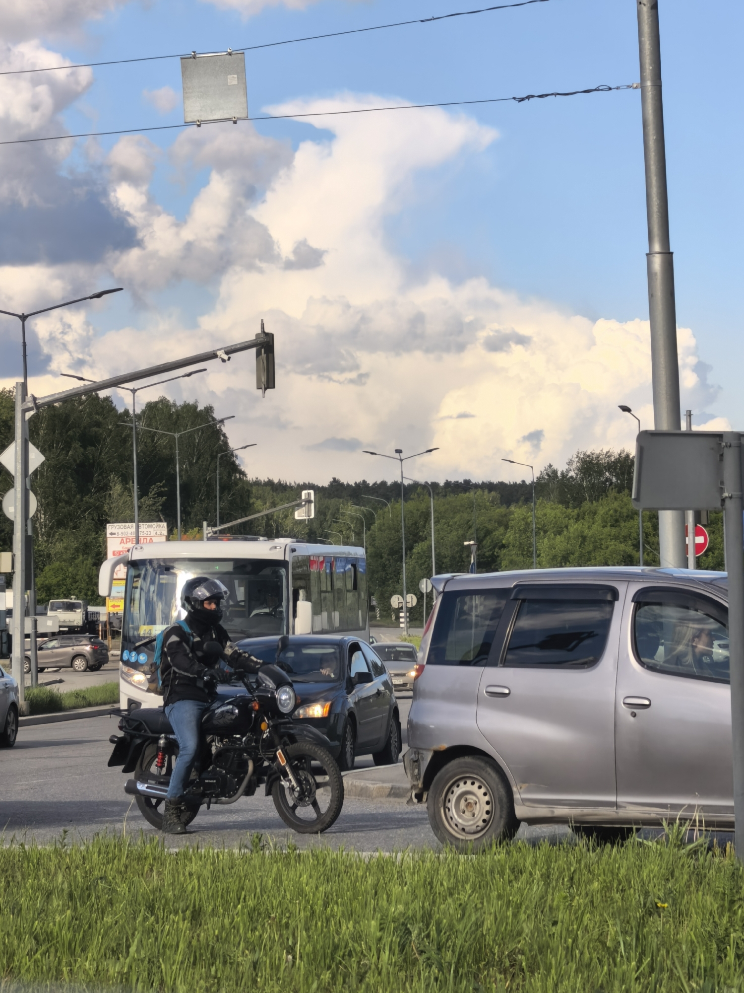The image shows a street scene in an urban environment. A motorcyclist, wearing a helmet and jacket, is stopped at a traffic light in the median lane. A silver van with a damaged bumper is to the right, partially obscured by a road sign. A bus and a dark-colored car are stopped behind the motorcyclist. A grassy median is in the foreground. Trees are visible in the background under a partly cloudy sky. Streetlights and overhead wires are present.