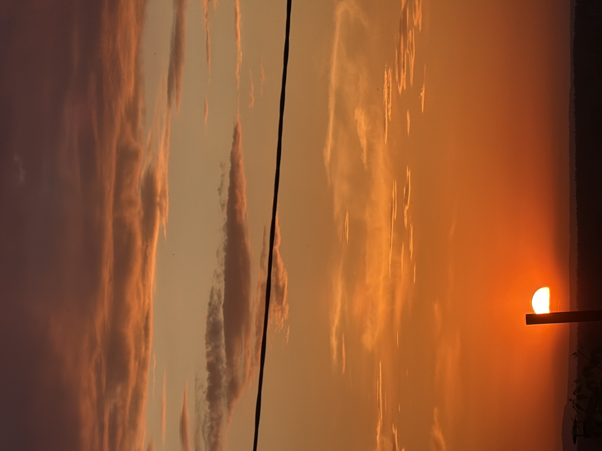 The image captures a sunset in what appears to be Chaikovsky, Russia. The sky is ablaze with orange and yellow hues, indicating the sun is low on the horizon. The sun is partially obscured by a horizontal surface, possibly a ledge or roof. Dark power lines cut through the sky on the left side of the frame. The clouds are streaky and dispersed. A dark treeline is visible on the right horizon.