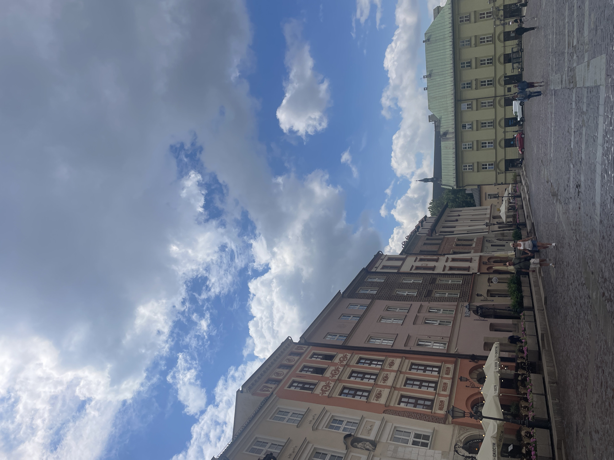 The image depicts a street scene in Krakow, Poland, likely within the Old Town. In the foreground, a cobblestone street stretches into the distance, lined with historic buildings on the left. These buildings feature ornate facades, with pastel colors like peach and light pink dominating some of the structures. Several of them have multiple stories with evenly spaced windows and decorative architectural elements. Outdoor seating with white umbrellas and tables are set up in front of some establishments, suggesting a cafe or restaurant setting. A few people are visible on the street.
Further down the street, on the right side of the image, a larger, light yellow building with a green roof stands prominently. This building also has many windows and appears to be a significant structure within the square or plaza. The sky above is a mix of blue and white clouds, with some larger, darker clouds indicating potential for varied weather.
In the background, a steeple or tower of a church or cathedral is visible, adding to the historical atmosphere of the location. Other vehicles, including a red car and a white van, are present in the middle ground. Several individuals are walking or standing in the plaza area in front of the green-roofed building, some appearing to be tourists. The overall impression is one of a vibrant historical city square on a day with dynamic cloud cover.
