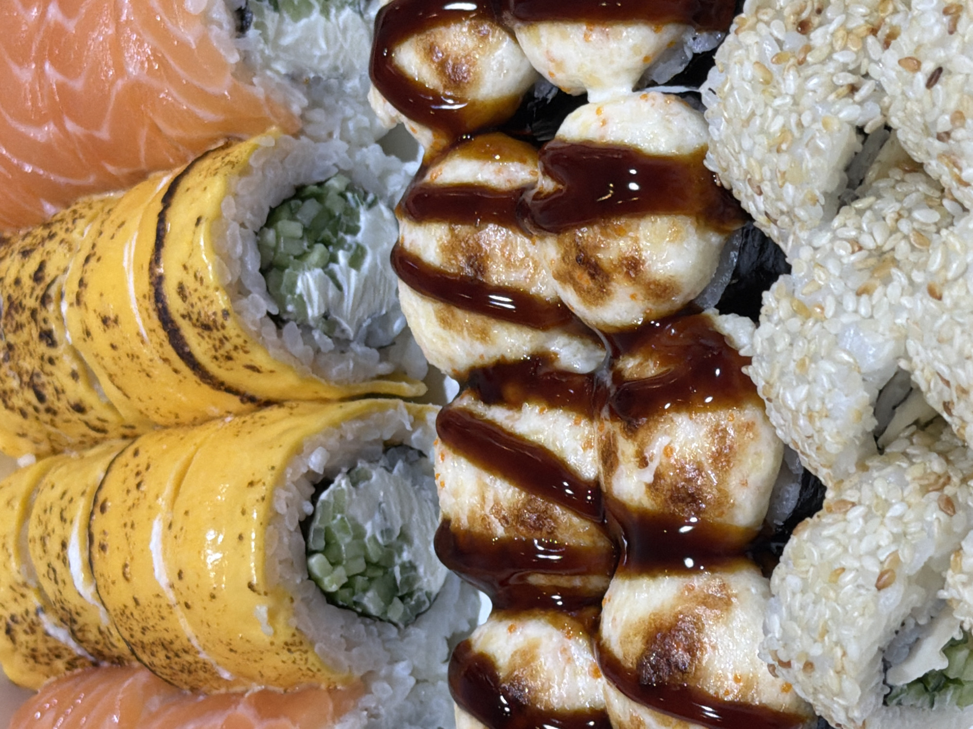 In Khmelnytskyi, Ukraine, a close-up, overhead shot displays a variety of sushi rolls arranged in a container. The sushi is artfully presented, showcasing different textures and colors. In the foreground, several rolls are covered in white sesame seeds. Behind them, another row of sushi is topped with a creamy, lightly browned sauce drizzled with a dark, glossy sauce, possibly teriyaki or eel sauce. Further back, rolls with a bright yellow topping, possibly omelet or a cheese-based sauce, are visible. On the far left and right edges, slices of raw salmon with their characteristic marbling are clearly discernible, indicating salmon nigiri or sashimi. The lighting is bright, highlighting the glossy sheen of the sauces and the fresh appearance of the ingredients. There are no people or other discernible environmental elements present in the frame, focusing solely on the food.