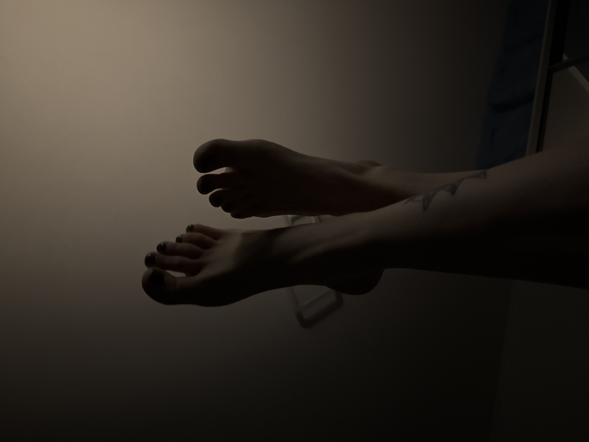 The image shows a pair of human feet and ankles against a dimly lit background. The left foot, with visible toes, is positioned closer to the bottom of the frame. The toes appear to have dark nail polish. The right foot is positioned slightly behind and above the left, with the toes curled inward. The ankles and part of the lower legs are visible, and there is what appears to be a tattoo on the visible part of the right leg. The background is mostly dark with faint light, and the upper right corner shows part of a light-colored wall with a blue object near the edge. The location is unknown; the image does not provide any information indicating it was taken in Moscow, Russia.