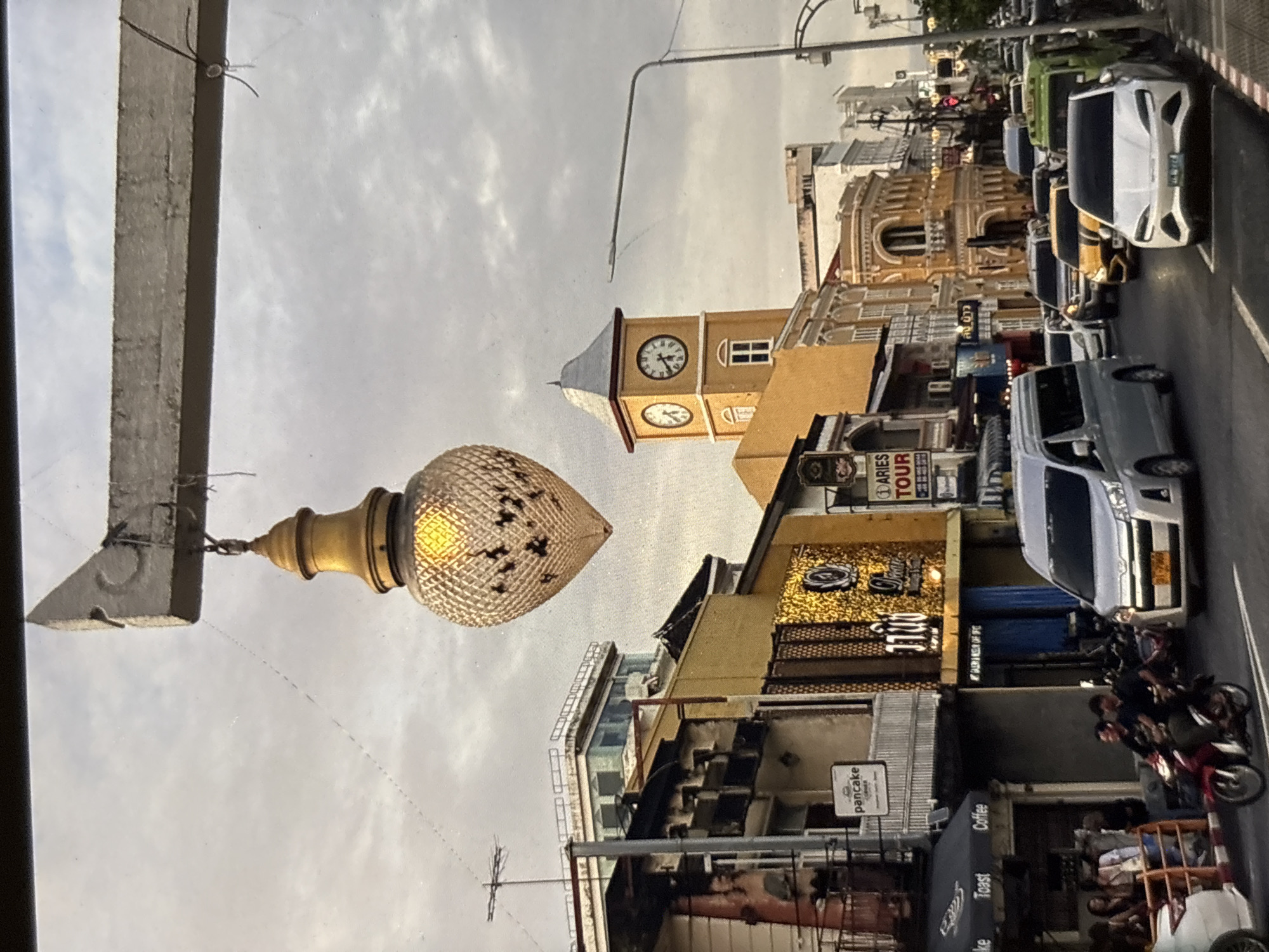 The image depicts a street scene in Chalong, Thailand, likely during the daytime or early evening. In the foreground, a vintage-style street lamp with a large, ornate, golden, heart-shaped glass shade hangs from a concrete beam. The street is bustling with traffic, including a silver minivan, a white sedan, a yellow car, and several other vehicles partially obscured by the camera's angle. A red motorcycle carrying two individuals is visible in the left lane.
The background features a row of buildings with distinct architectural styles. The building directly behind the motorcycle has a golden facade with intricate patterns. To the right of this building, a prominent clock tower with two visible clock faces stands tall, painted in yellow and white. Further down the street, more buildings are visible, some with ornate balconies and arches. A street sign with the word "pancake" is affixed to one of the buildings on the left. The sky is overcast, suggesting cloudy weather. The overall atmosphere appears to be that of a lively urban environment.