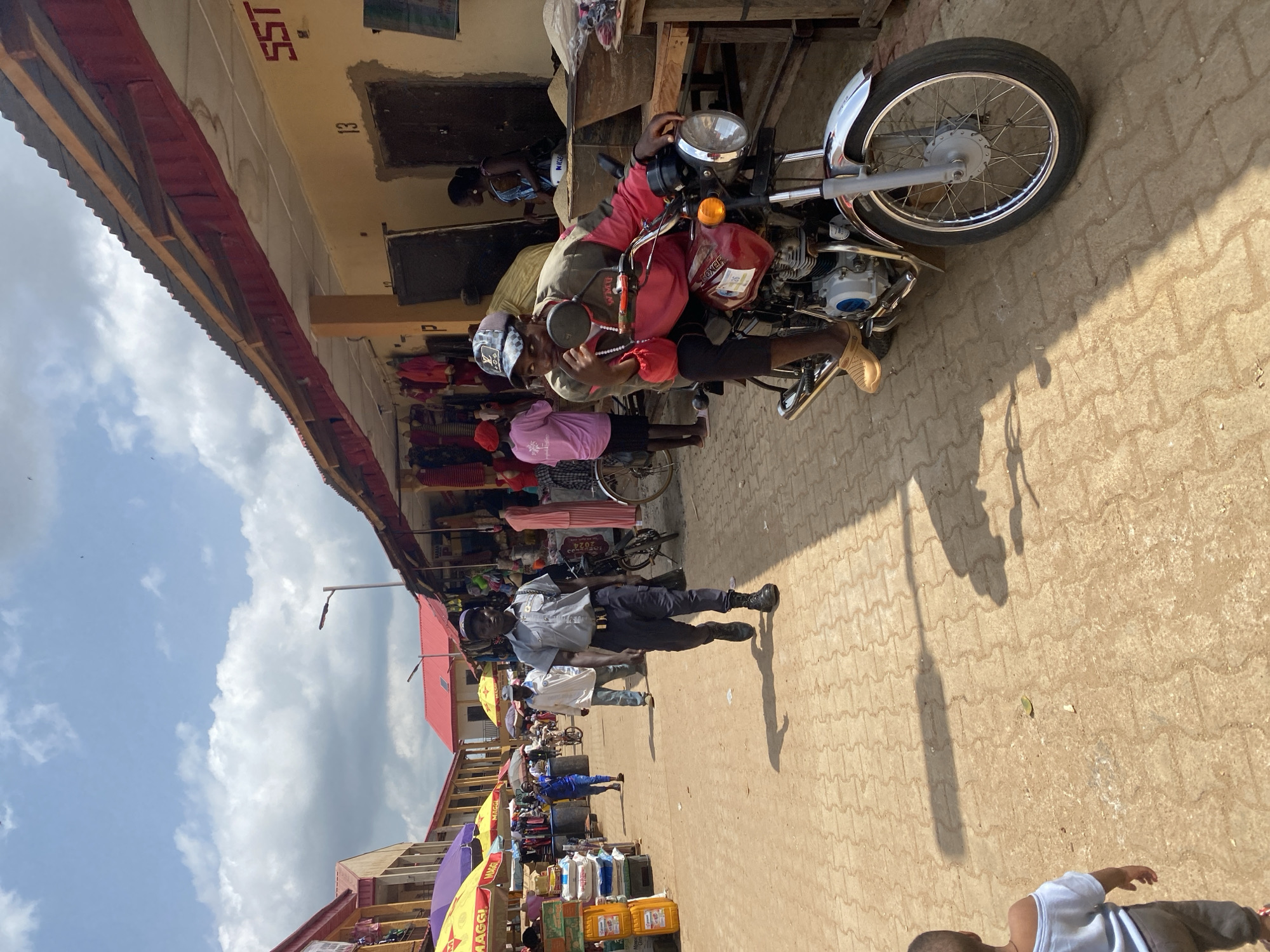This is a street-level photograph taken in Kafanchan B, Nigeria. The scene appears to be a busy marketplace with stalls and a street. The photograph is oriented vertically, showing a street paved with interlocking bricks.
The primary subjects are people and objects. In the foreground, a man in a grey uniform and dark trousers is walking on the street, casting a long shadow. He is wearing a cap and boots. Slightly behind him, another man in a white shirt and jeans is visible. Further down the street, several people are browsing or working at market stalls, which display various goods including clothing and packaged items. A red motorcycle is prominently featured in the middle ground, with a person seated on it, wearing a red jacket and a cap.
The activity appears to be a typical day at a marketplace, with people engaged in commerce and movement. The time of day is likely daytime, indicated by the bright sunlight and the long shadows cast by the people and objects. The weather seems fair, with a mix of blue sky and clouds visible in the upper portion of the image.
Notable details include the presence of market stalls with awnings, suggesting a commercial hub. The motorcycle and the variety of goods on display provide context for the location and its activities. The emotion conveyed is one of everyday life and activity.
Visible text includes "SST" on a building in the background, "13" next to it, and "MAGGI" on some yellow packaging. There is also a label on the red motorcycle that appears to say "BOXER".
