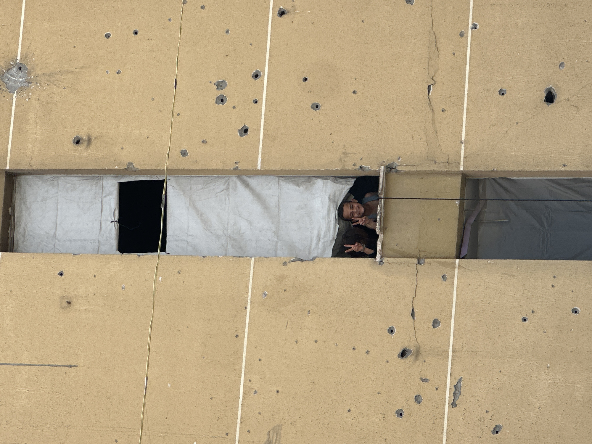 The image shows the exterior of a building in Unknown, Unknown, that appears to have sustained damage, possibly from gunfire or explosions, judging by the numerous holes in the walls. A person is visible in an opening in the wall, making a peace sign with both hands. The opening is partly covered with what appears to be fabric or sheeting.