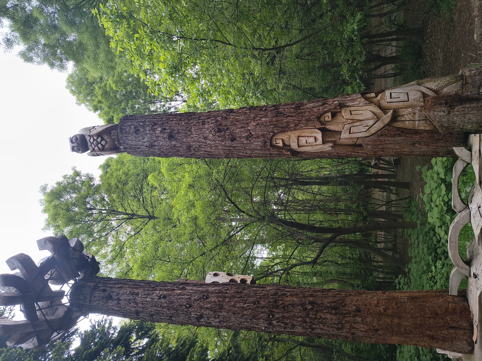 The image shows two wooden carvings in a wooded area. The carving on the left has a wind vane at the top, while the carving on the right has an owl perched on top and carvings of geometric shapes. The location is in the city of Unknown, Unknown. The environment consists of green trees, a wooden bench, and a path.