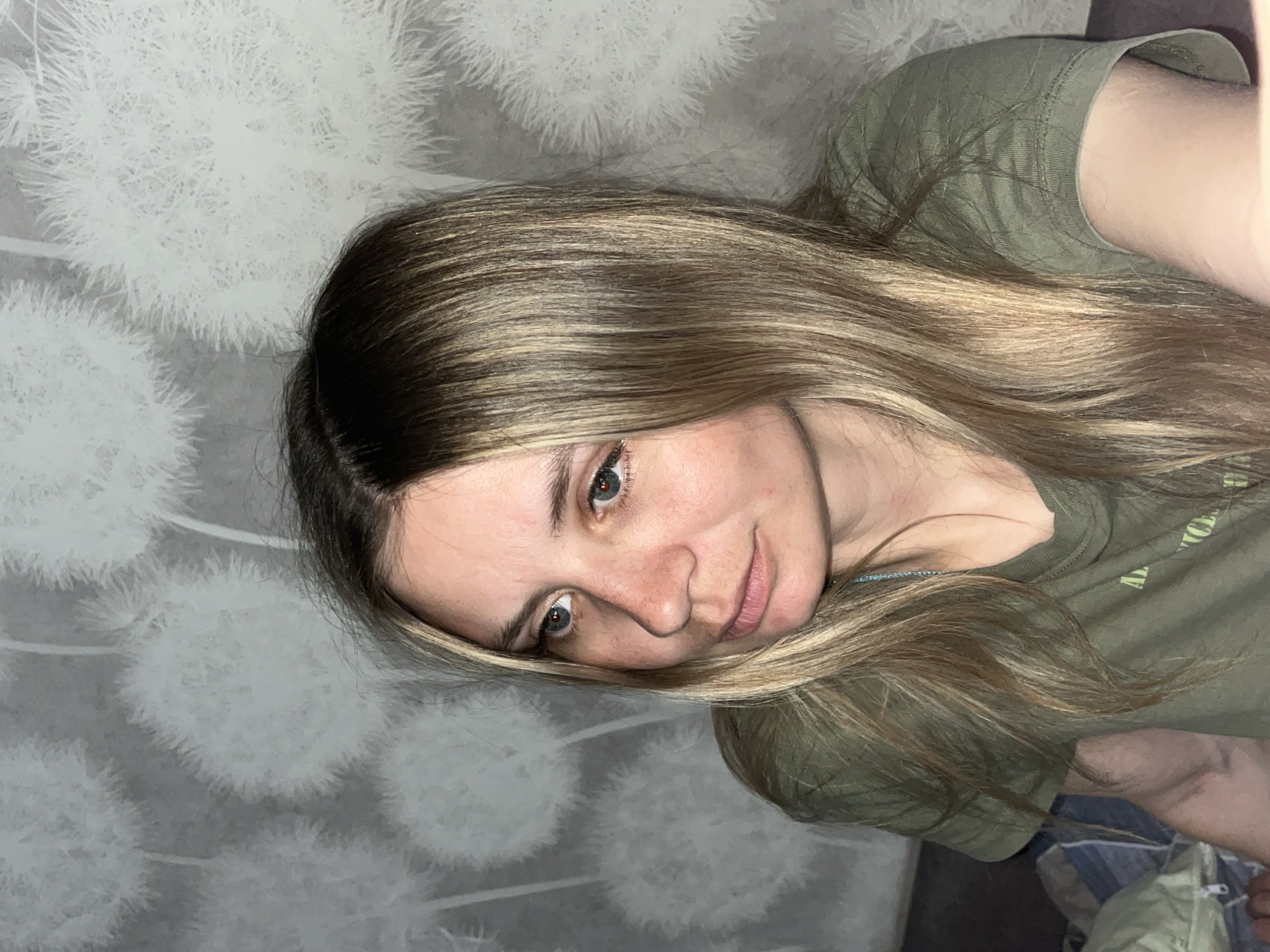 A woman with long, blonde highlighted hair is lying on a light gray patterned surface. She is wearing an olive green t-shirt. The background features a light gray wall with a dandelion-like pattern. The photograph appears to have been taken indoors. No specific elements indicate the location to be in Chaikovsky, Russia.