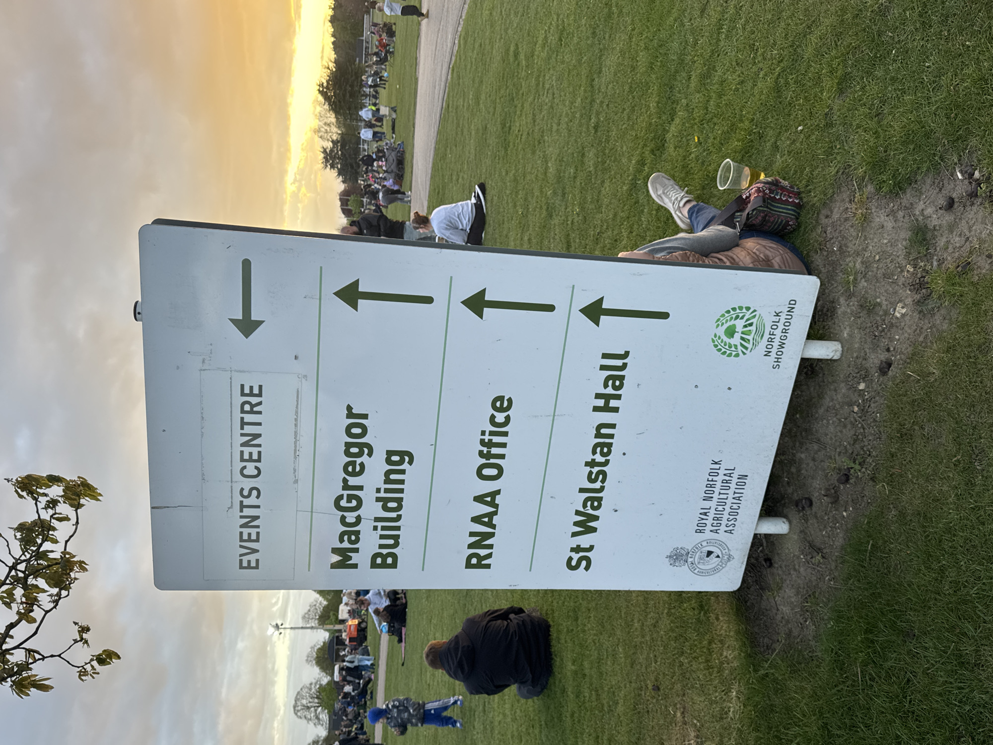 The image captures a scene outdoors, likely during an event at the Royal Norfolk Showground, South Norfolk, United Kingdom. The foreground is dominated by a signpost indicating directions to the Events Centre, MacGregor Building, RNAA Office, and St Walstan Hall. A person is lying down next to the sign. In the background, there's a gathering of people on a grassy area, suggesting an ongoing event. The sky above indicates the time to be late afternoon or early evening. The overall atmosphere suggests a relaxed, outdoor event setting.