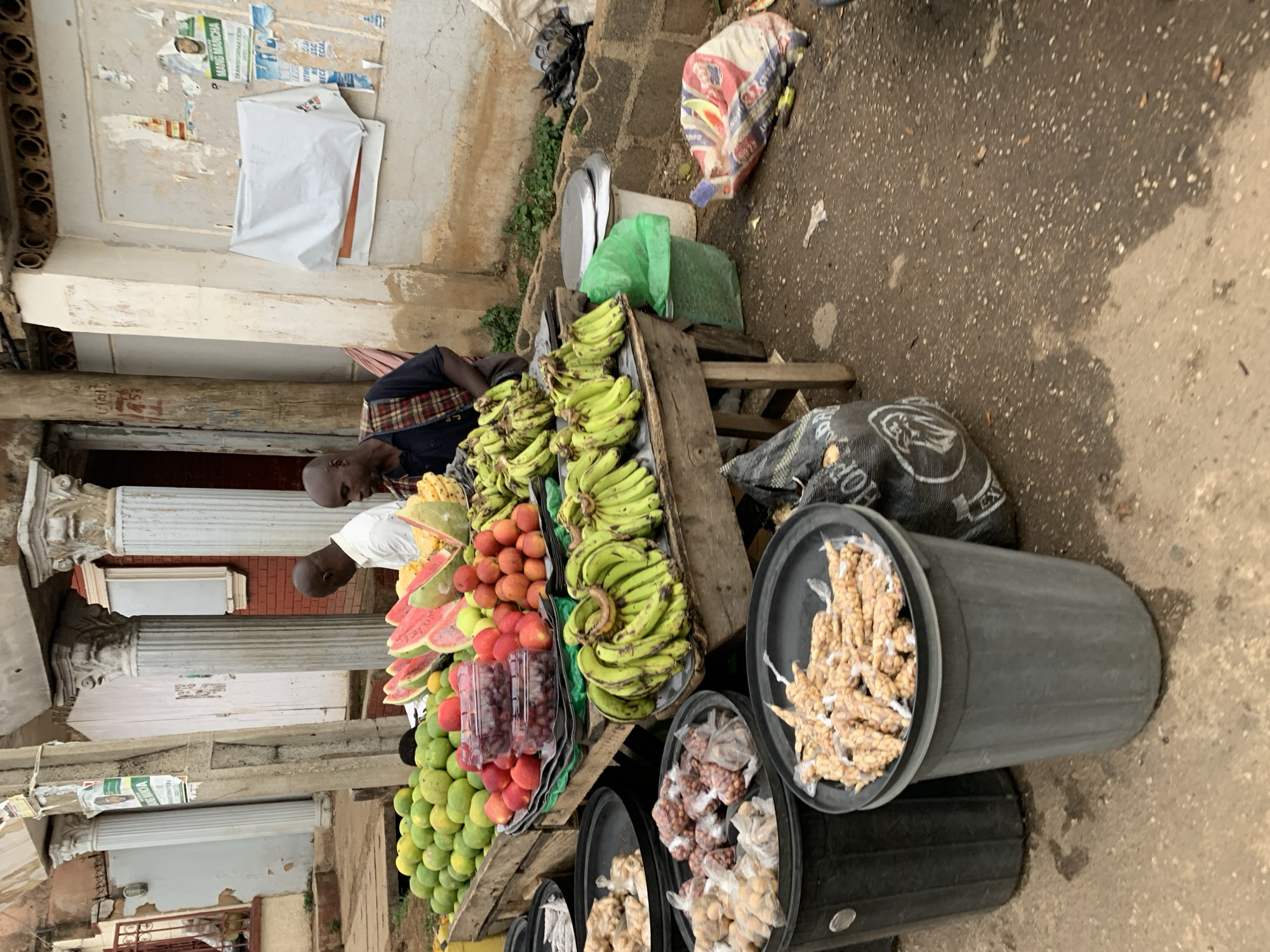 In an outdoor market at New Stadium, Nigeria, two men are tending to a stall laden with fruits and snacks. The stall displays a variety of produce, including bunches of green bananas, watermelons, apples, plums, and mangoes. Two black plastic bins contain individually wrapped snacks, likely peanuts or other nuts. A weighing scale and a green plastic bag are positioned near the bananas. The stall is located in front of a building with classical architectural details, including columns. The street is paved.