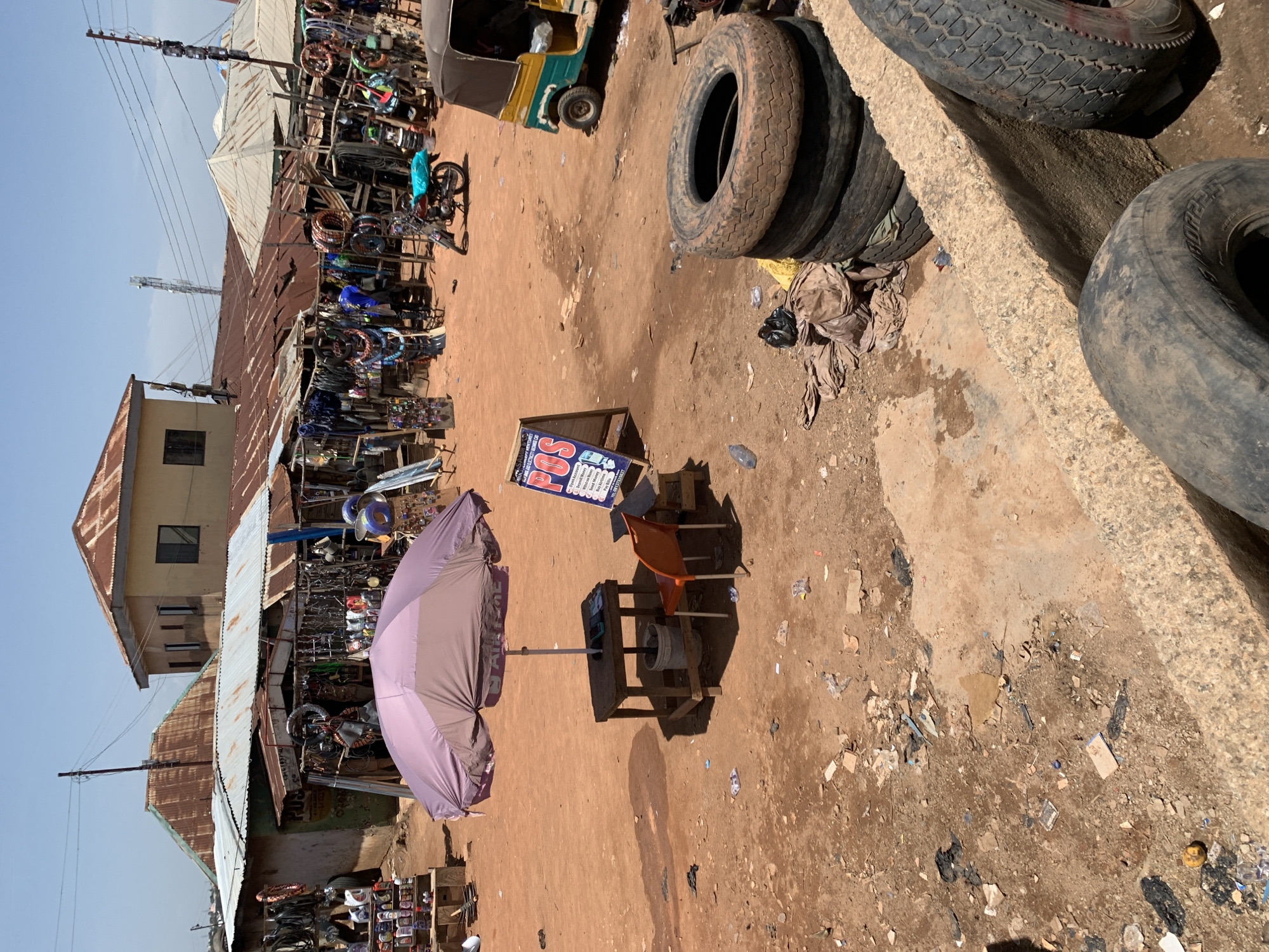 This image depicts an outdoor market or commercial area in Gura Top, Nigeria, under bright, clear skies. The ground is a reddish-brown, dusty dirt, littered with various debris including plastic bottles, paper, and fabric scraps.
In the immediate foreground and extending to the right, several large, worn vehicle tires are stacked against a rough concrete barrier. In the mid-ground, a purple parasol, possibly bearing the logo "AIRTEL," provides shade over a simple wooden table and an orange plastic chair. On the table, a bucket or container is visible.
Adjacent to the table, a blue and white sign prominently displays "POS" in red letters. Below, smaller text lists financial services offered, including "Withdrawal," "Deposit," "Transfer," "Airtime Top-up," "Data," "Bills Payment," and "Registration," along with a phone number.
Further into the background and to the left, a row of market stalls with corrugated metal roofs lines the street. These stalls appear to sell various goods, prominently featuring numerous bicycle and motorcycle parts, such as rims, tires, and other metallic components. A green and yellow auto-rickshaw is parked in the middle-ground, and another motorcycle is visible near the stalls. Beyond the market stalls, a few buildings with corrugated roofs are visible, including a multi-story structure. Utility poles with numerous power lines crisscross the upper left portion of the frame.
The strong shadows suggest it is midday, with direct sunlight. No specific human activity is clearly visible in detail, but the setup indicates an active commercial environment.