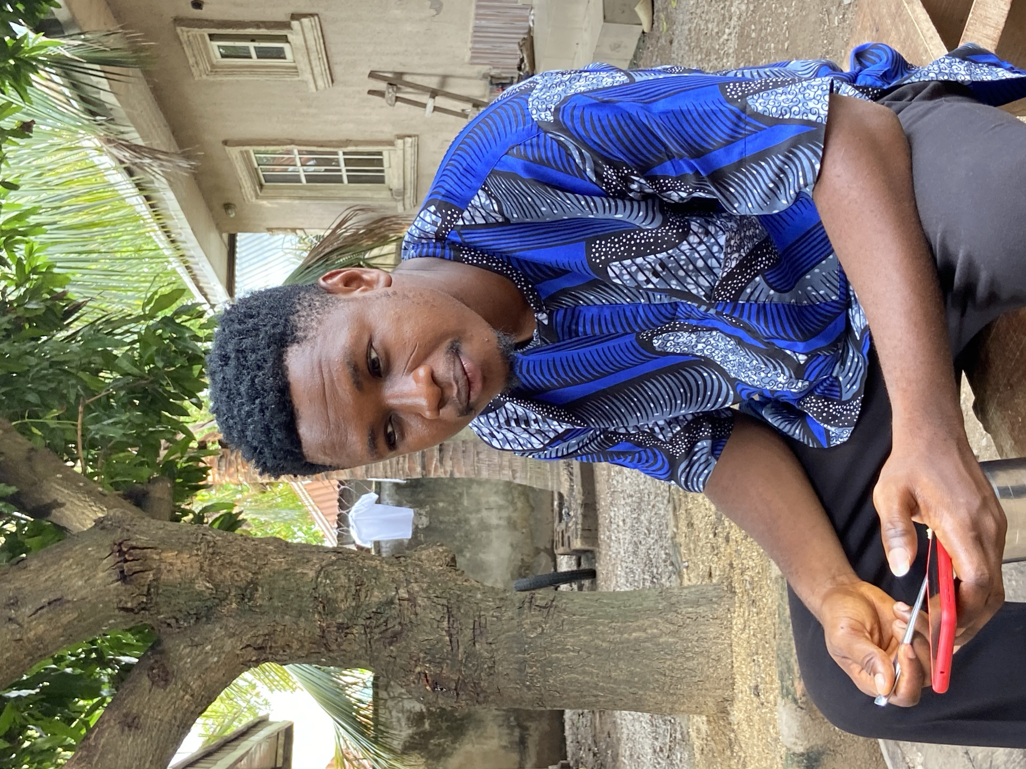 A man sits outdoors in Mararaba, Nigeria. He is wearing a blue patterned shirt and black pants, holding a red and silver tool in his hands. A tree is to his left, with a white cloth hanging in the background. A building with windows is visible behind the tree.