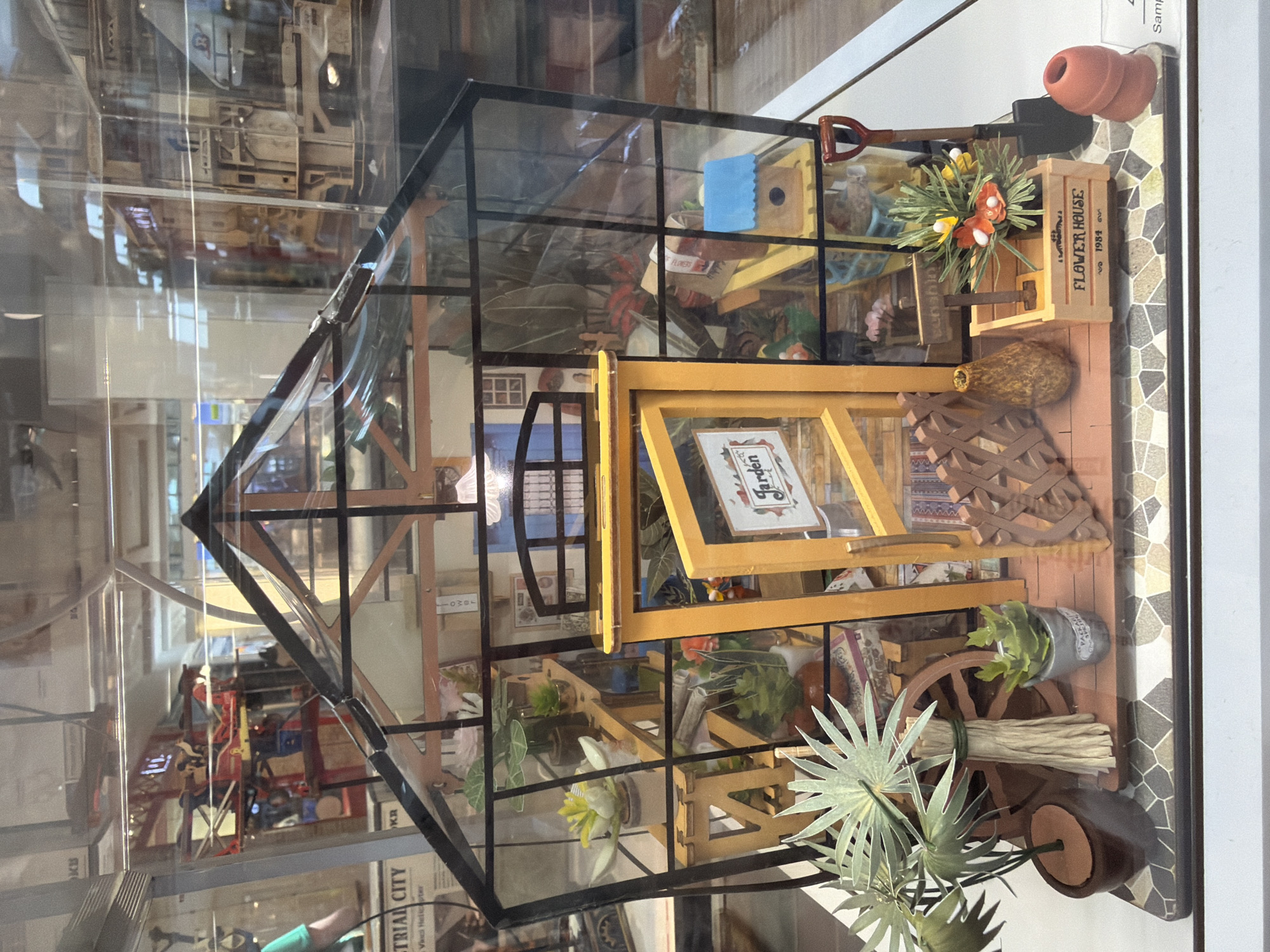 This is a top-down, eye-level, medium shot of a miniature greenhouse display, viewed through glass, possibly in a museum or store. The greenhouse itself is made of clear acrylic panels with black framing, and it is filled with various miniature gardening-related items.
Inside the greenhouse, there are shelves lined with tiny potted plants, some with vibrant green leaves and others with delicate white and yellow flowers. A miniature wooden crate labeled "FLOWER HOUSE" sits on a shelf, holding a bouquet of artificial orange and yellow flowers. A small toy shovel rests on top of the greenhouse, and a tiny birdhouse with a blue decorative element is also visible. Other details include a small wooden wagon wheel, bundles of straw, and various decorative elements that mimic gardening tools and accessories.
In the background, reflections show what appears to be a larger display or exhibit, with more complex structures and objects, possibly representing a city or industrial scene. The lighting is bright and appears to be artificial, likely from indoor display lighting. The overall scene evokes a sense of intricate craftsmanship and a miniature world dedicated to gardening. There is no visible text on the greenhouse itself, but the "FLOWER HOUSE" label on the crate is legible. The reflections in the glass obscure some details of the background display.