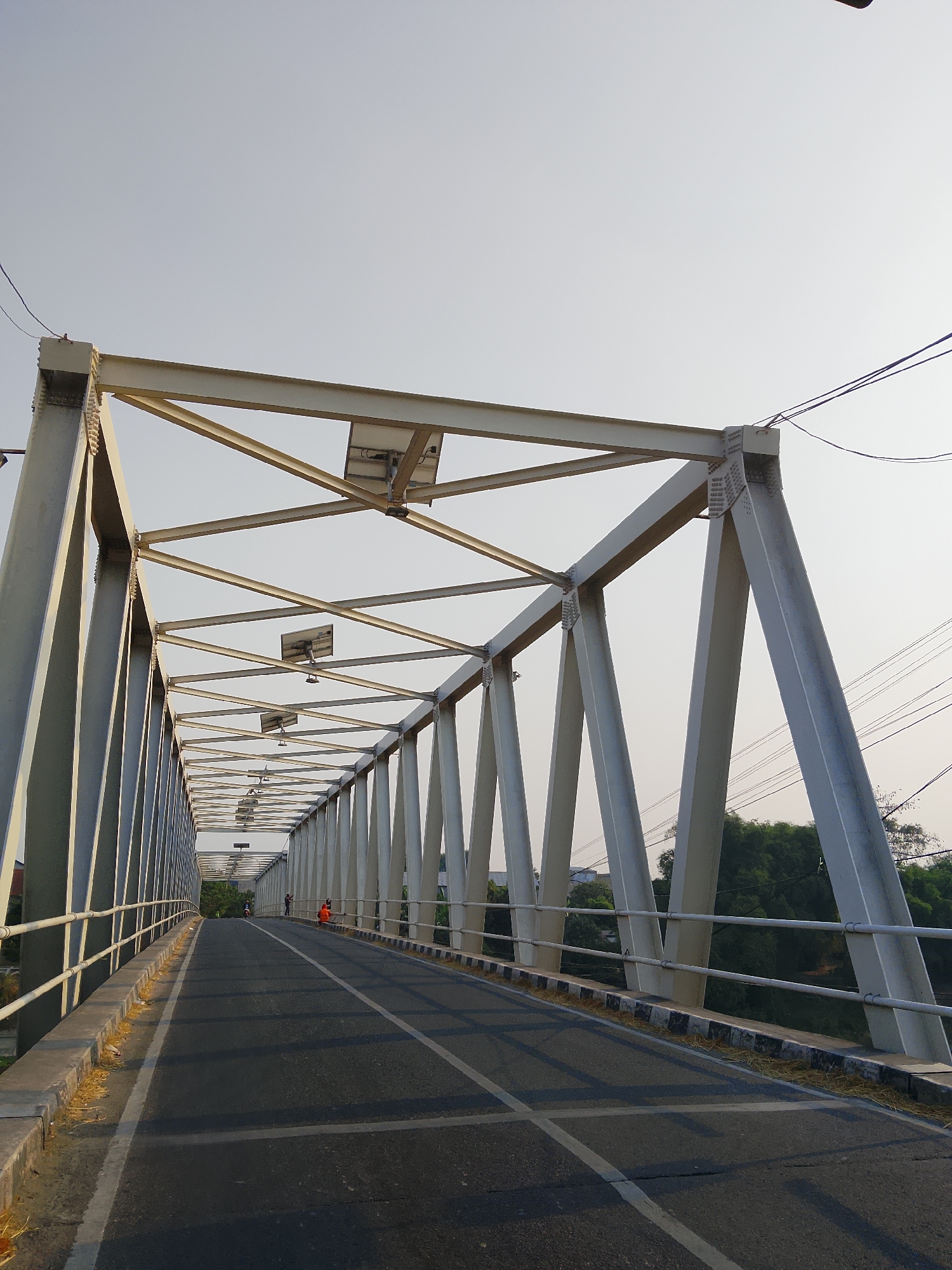The image shows a perspective view from a road onto a white truss bridge spanning a body of water. The bridge has a clear roadway with lane markings, bordered by white railings. Along the top of the bridge's structure, solar panels are visible. In the distance, on the bridge, two individuals can be seen, one wearing an orange vest, and another person appears to be on a motorcycle. The surrounding environment includes dense green trees and foliage on the banks of the waterway. The sky is clear and pale. The scene is indicative of the Kasiman bridge in Kasiman, Indonesia, a structure that facilitates transportation over a river or canal in a semi-urban or suburban setting.