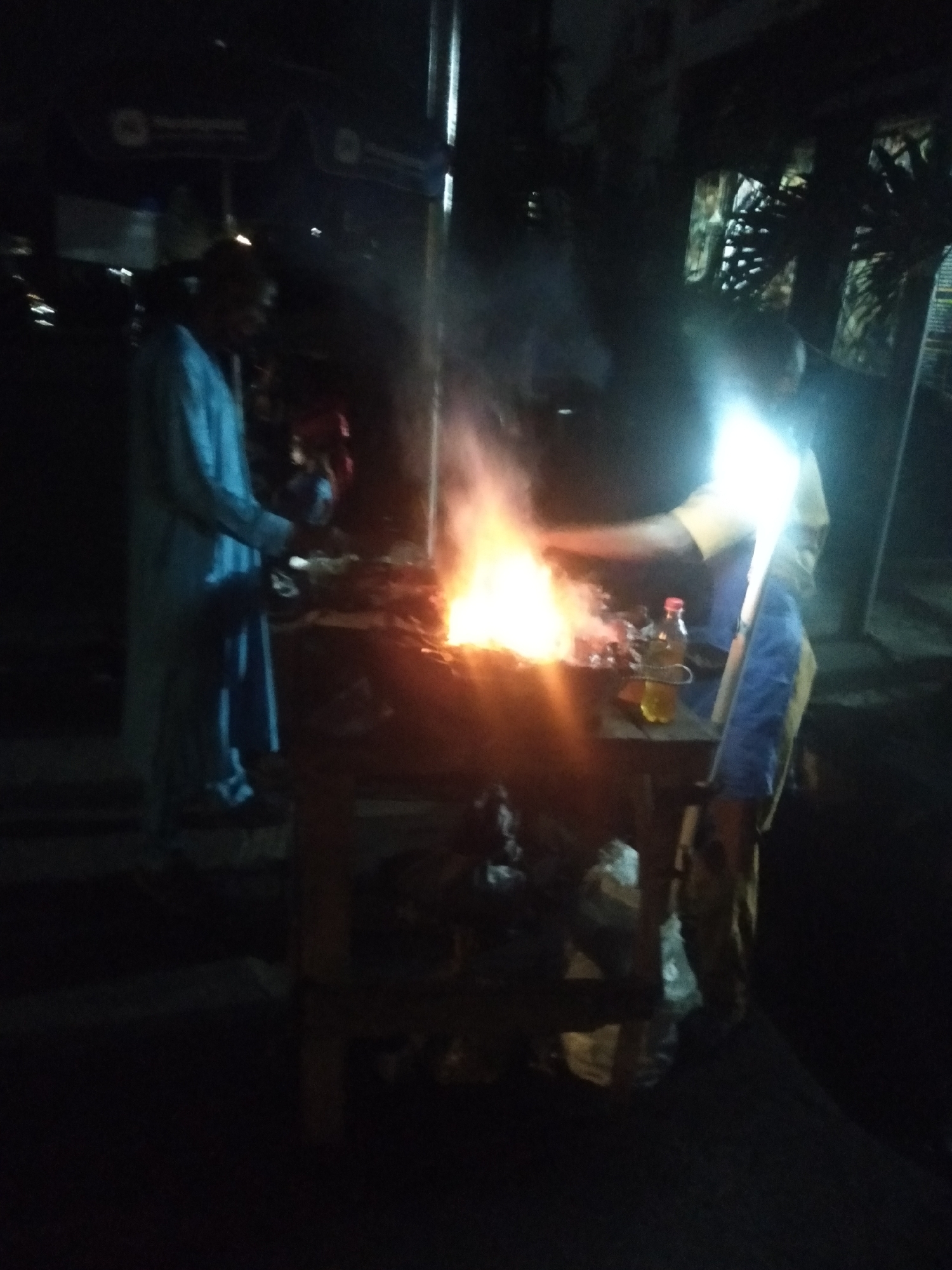 This nighttime image captures a street food vending scene, likely in Shangisha, Nigeria. Two individuals are present, engaged in cooking over an open fire.
At the heart of the scene is a bright, orange-yellow fire, burning on a makeshift wooden table or stand. Smoke rises visibly from the flames, beneath which dark, irregularly shaped items (suggesting grilled food like suya) are being cooked. A clear plastic bottle with a red cap, containing a yellowish liquid (likely cooking oil), rests on the table close to the fire.
On the right side of the fire, a person in a light-colored top (possibly yellow or beige) and blue trousers or an apron is actively tending to the cooking. A powerful, bright light source, appearing to be attached to their upper body, illuminates them intensely, creating a strong lens flare and obscuring their facial features.
To the left of the fire, another person stands, clad in a long, light-colored, possibly blue or light grey, traditional garment. Their face is largely obscured by the darkness of the night, and they appear to be observing or assisting with the preparation.
The background is dark and blurry, typical of a low-light environment. Faint outlines of structures or vehicles are visible on the left, along with a barely discernible sign or banner that appears to have a circular logo. A vertical pole is visible on the far right. The image conveys the atmosphere of a bustling night market or roadside food stall, with the fire serving as both a cooking implement and a primary source of light.