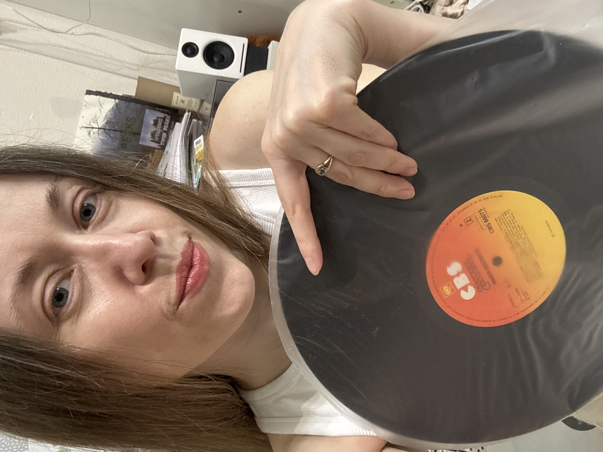 A close-up, selfie-style image captures a woman holding a black vinyl record in an indoor setting, presumably in Kudrovo, Russia. The woman, with long brown hair and light-colored eyes, looks directly at the camera with a slight, playful smirk, her lips accentuated by a reddish gloss. She is wearing a white ribbed tank top and a gold ring with a blue stone on her right ring finger.
She holds the large vinyl record with both hands, partially encased in a clear protective sleeve. The record's central label is a vibrant orange and yellow gradient, prominently displaying the "CBS" logo in white. Visible text on the label includes "CBS 86075", "STEREO", "33 1/3 RPM", and "MADE IN HOLLAND". The OCR further identifies the word "Brothers" on the label.
In the background, a white cube-shaped speaker with two black circular drivers is visible on a surface. Behind the speaker and to the woman's left, a stack of books and papers is present. A book cover clearly displays the title "WELCOME TO TWIN PEAKS" with a scenic image of mountains and trees, and another book nearby shows the partial word "PEAKS", indicating its connection to the series. The background wall is plain and light-colored, with some white cables running along its upper edge. The indoor lighting does not indicate a specific time of day. The woman's direct gaze and expression convey a sense of engagement.