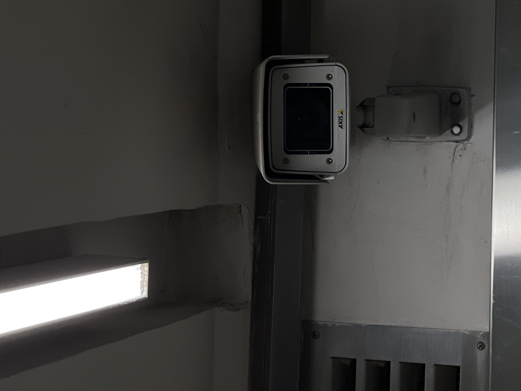 The image shows a surveillance camera mounted on a wall next to a vertical support beam. The camera is white with a black lens and the word "AXIS" printed on its side. It is attached to the wall via a white mounting bracket. Below the camera, the wall has a metal panel with horizontal slits. To the left of the camera, there's a horizontal light source. The location is not identifiable from the image.